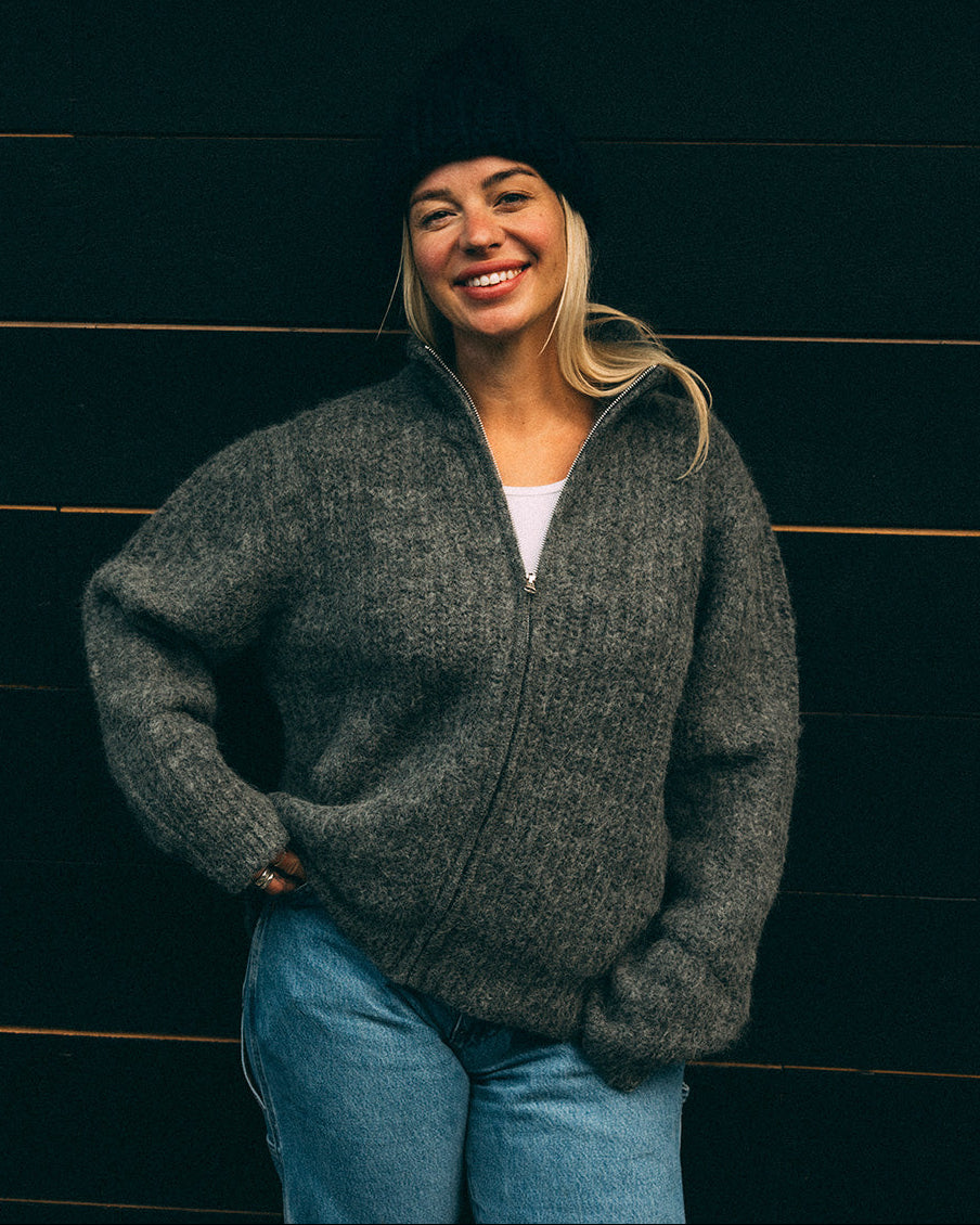 Woman wearing a grey Bare Knitwear full zip sweater and blue jeans against a dark wooden background