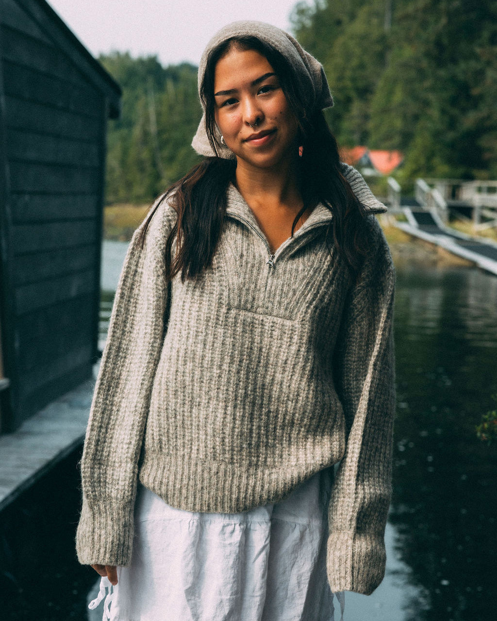 Woman wearing a beige Bare Knitwear 1/4 zip sweater and white skirt standing by a body of water with trees in the background.