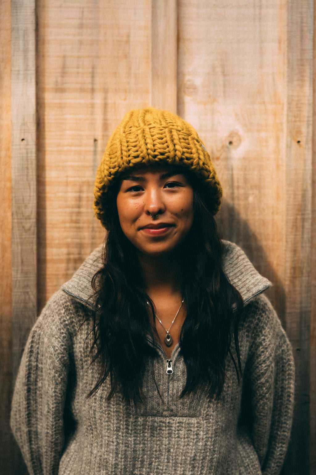 Person wearing a yellow diane rudge knit hat and gray Bare knitwear sweater against a wooden background. She is also wearing a Sow Song Stone necklace