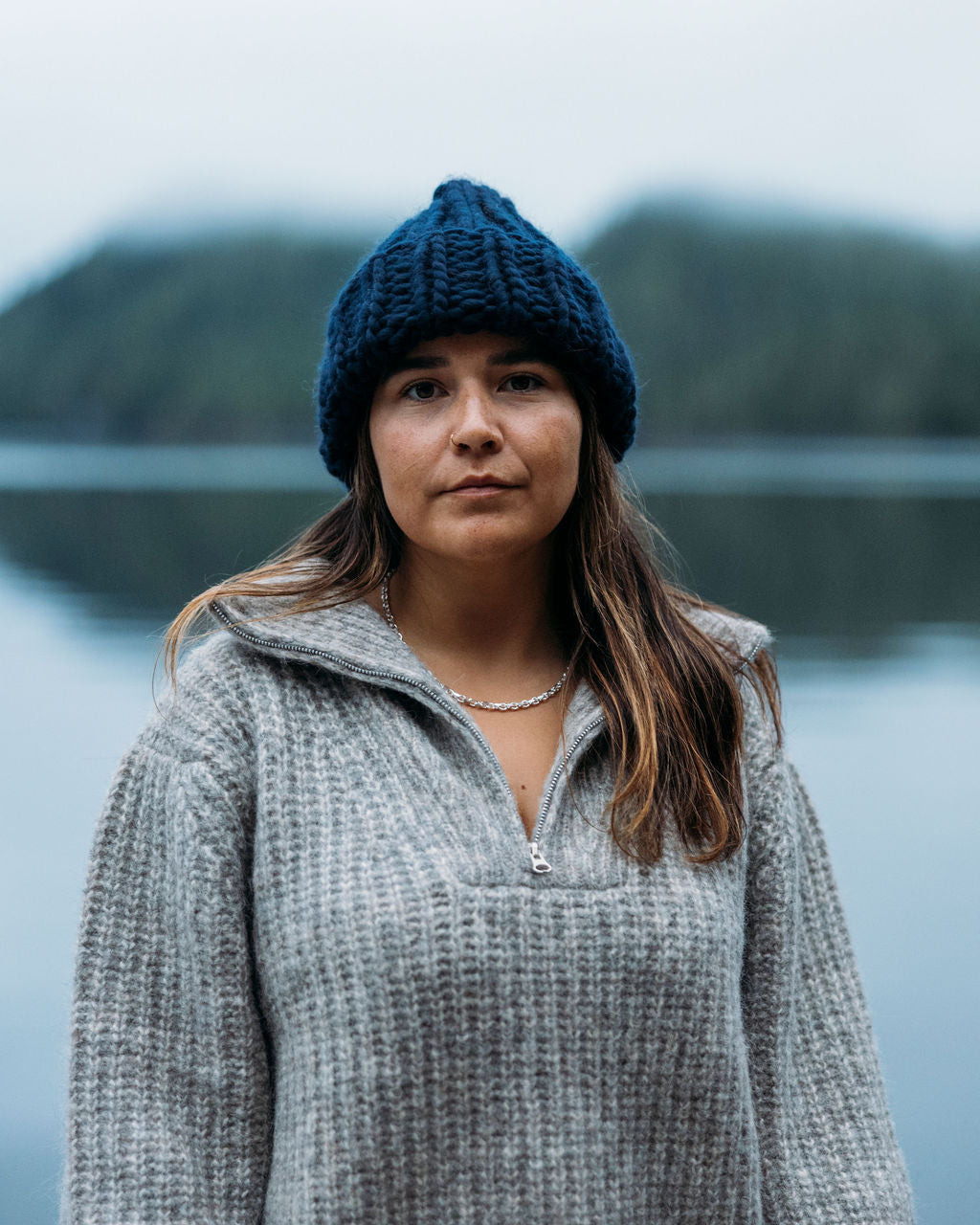 Woman wearing a blue Diane Rudge knit beanie, gray Bare Knitwear sweater and a Sow Song sterling silver chain standing in front of a blurred natural background