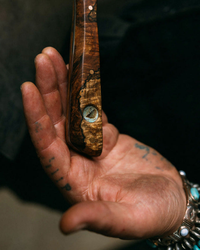 Hand holding a wooden knife handle with an abalone inalay against a dark background