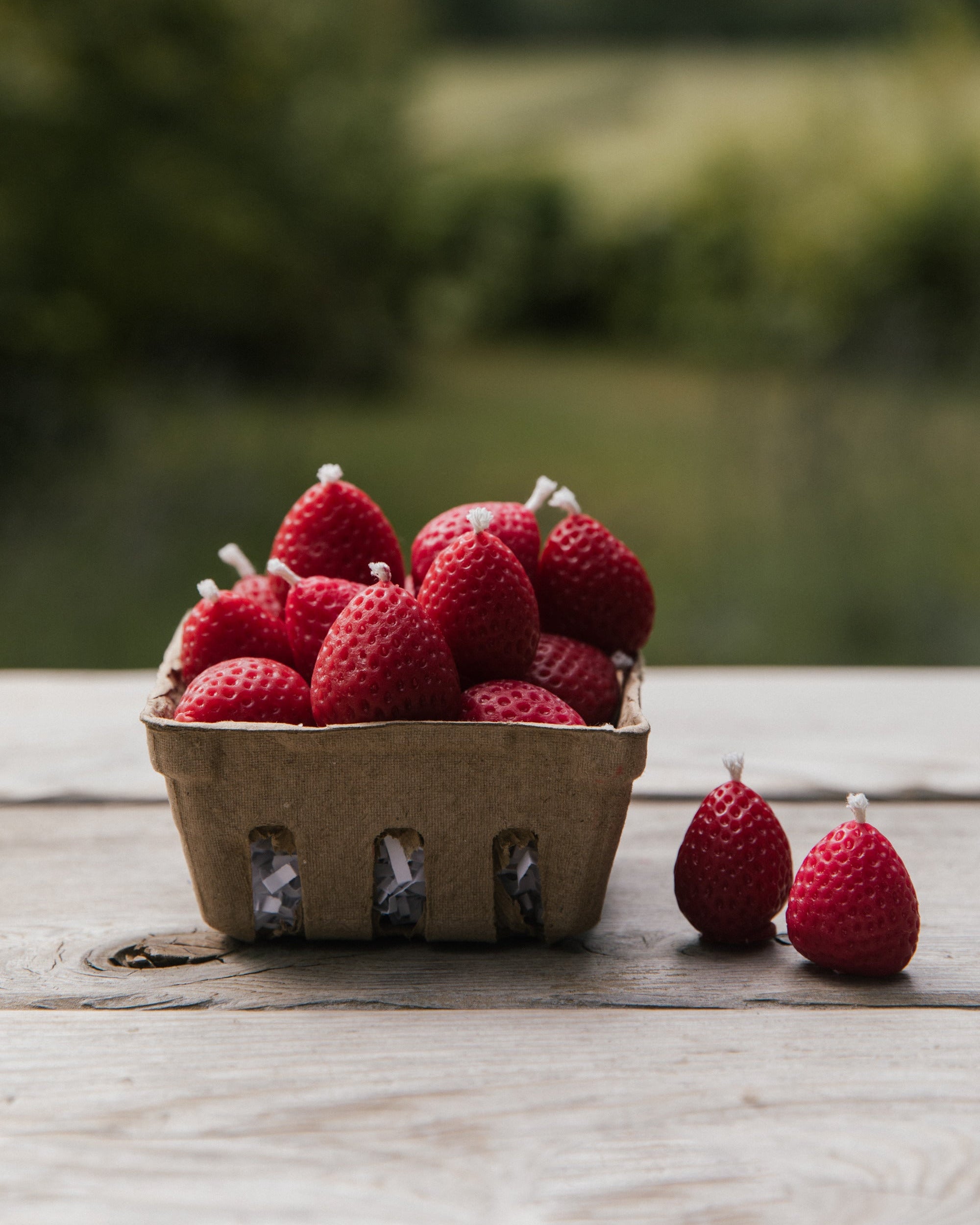 Strawberry candles in a cardboard container on a wooden surface with a blurred natural background