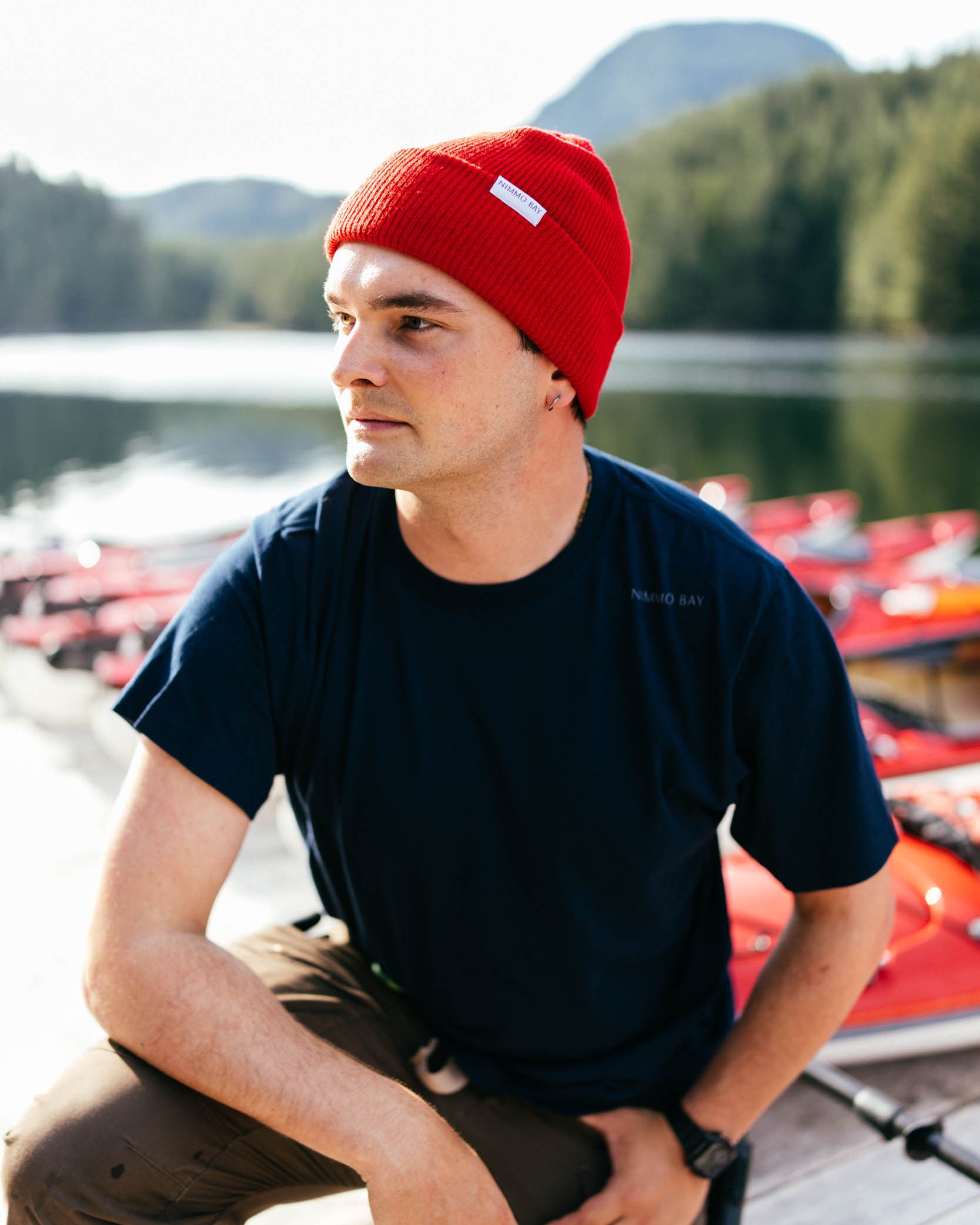 Man wearing a red beanie and navy shirt sitting on a dock with boats and water in the background