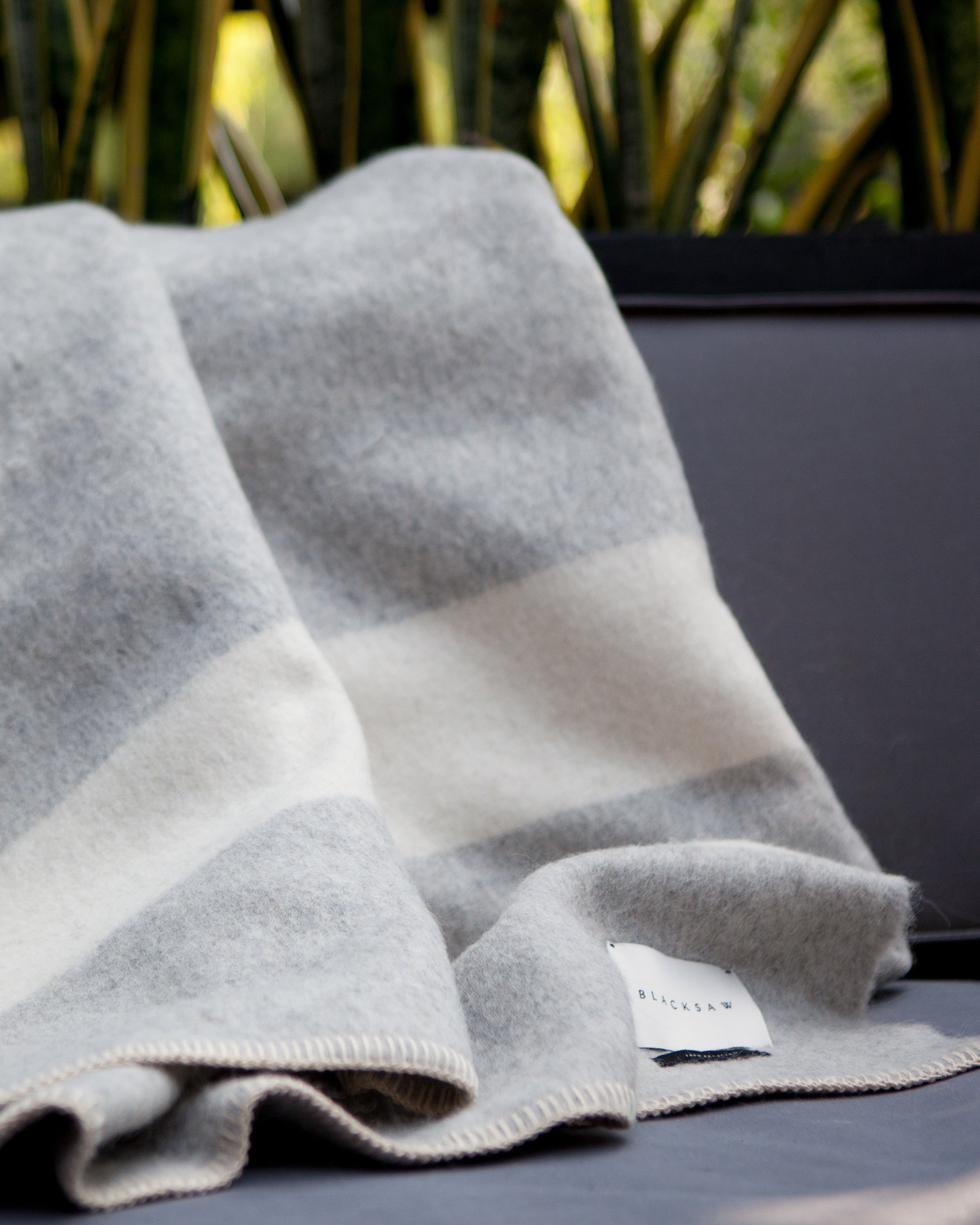 A folded light gray and cream striped wool blanket draped over a dark gray outdoor sofa, with blurred green foliage in the background.