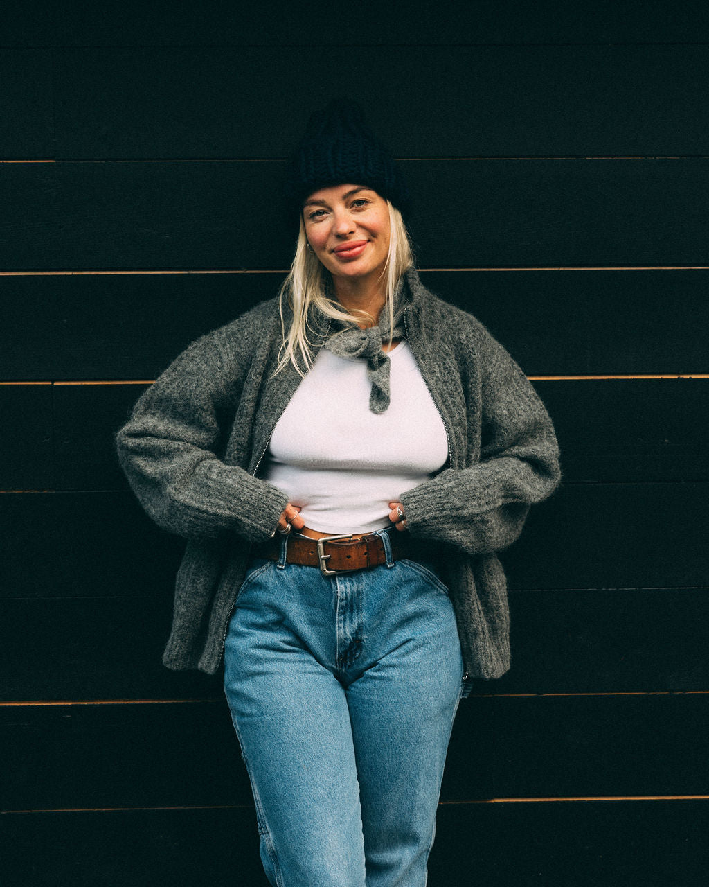 Woman wearing a grey Bare Knitwear full zip sweater, white shirt, blue jeans, and navy beanie against a dark wooden wall.