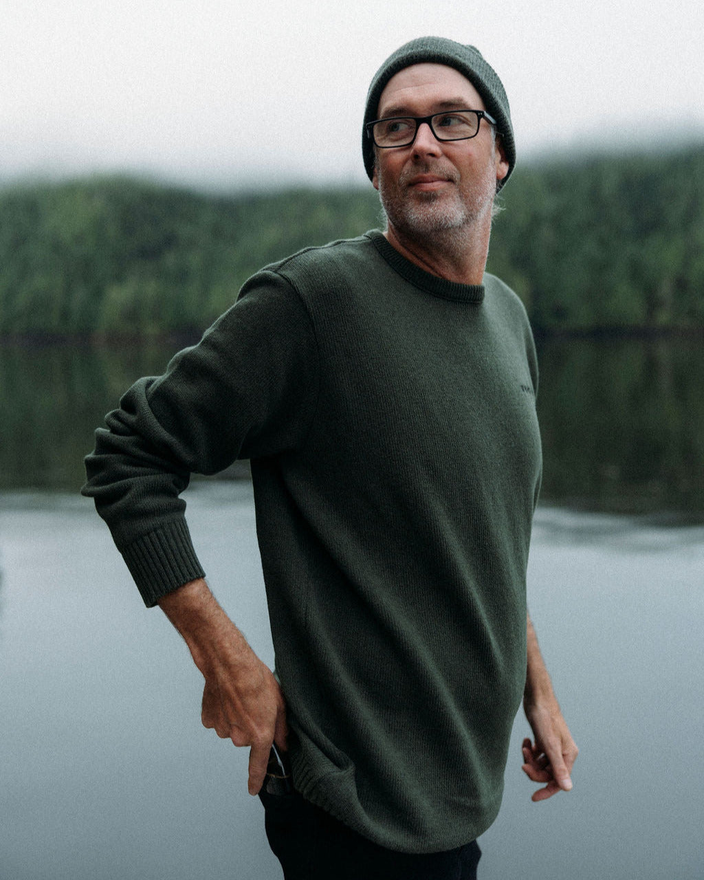 Man wearing a green anian fishermans sweater and beanie standing by a body of water with trees in the background