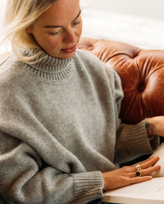 Woman reading a book wearing a The Heritage Turtleneck on a brown leather couch