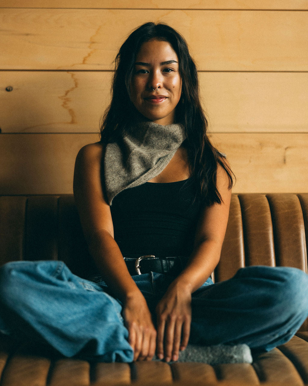 Woman sitting on a couch wearing a black top and blue pants with a bare knitwear bandana, wooden wall background