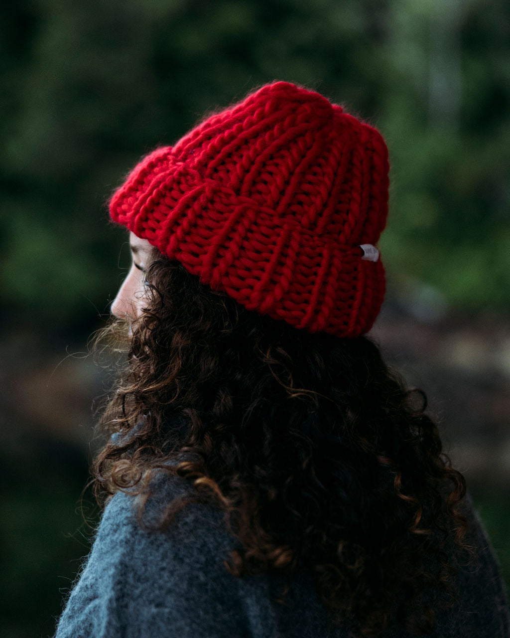 Person wearing a red Diane Rudge knit toque and grey Bare Knitwear sweater standing against blurred natural background