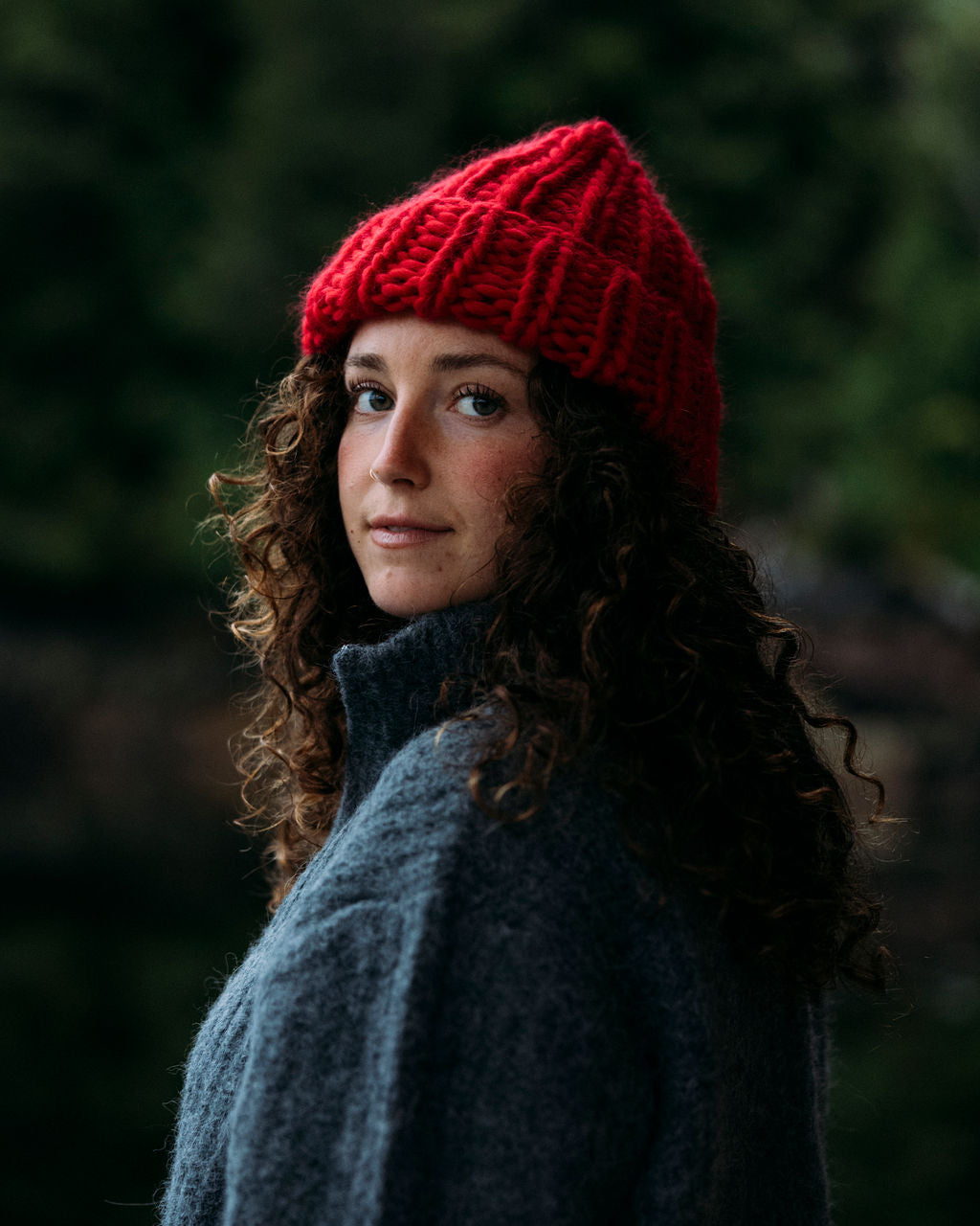 Person wearing a Red Diane Rudge knit toque and grey Bare Knitwear sweater standing against a forest background