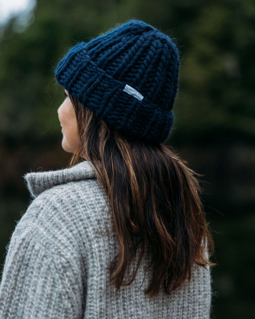 Person wearing a Navy Diane Rudge knit toque and grey Bare Knitwear sweater standing against a forest background