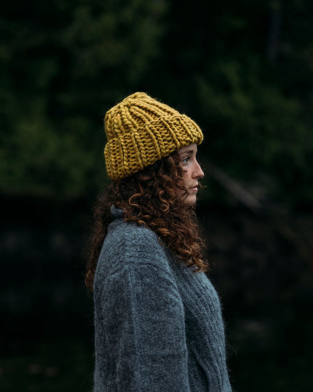 Person wearing a Ochre Diane Rudge knit toque and grey Bare Knitwear sweater standing against a forest background