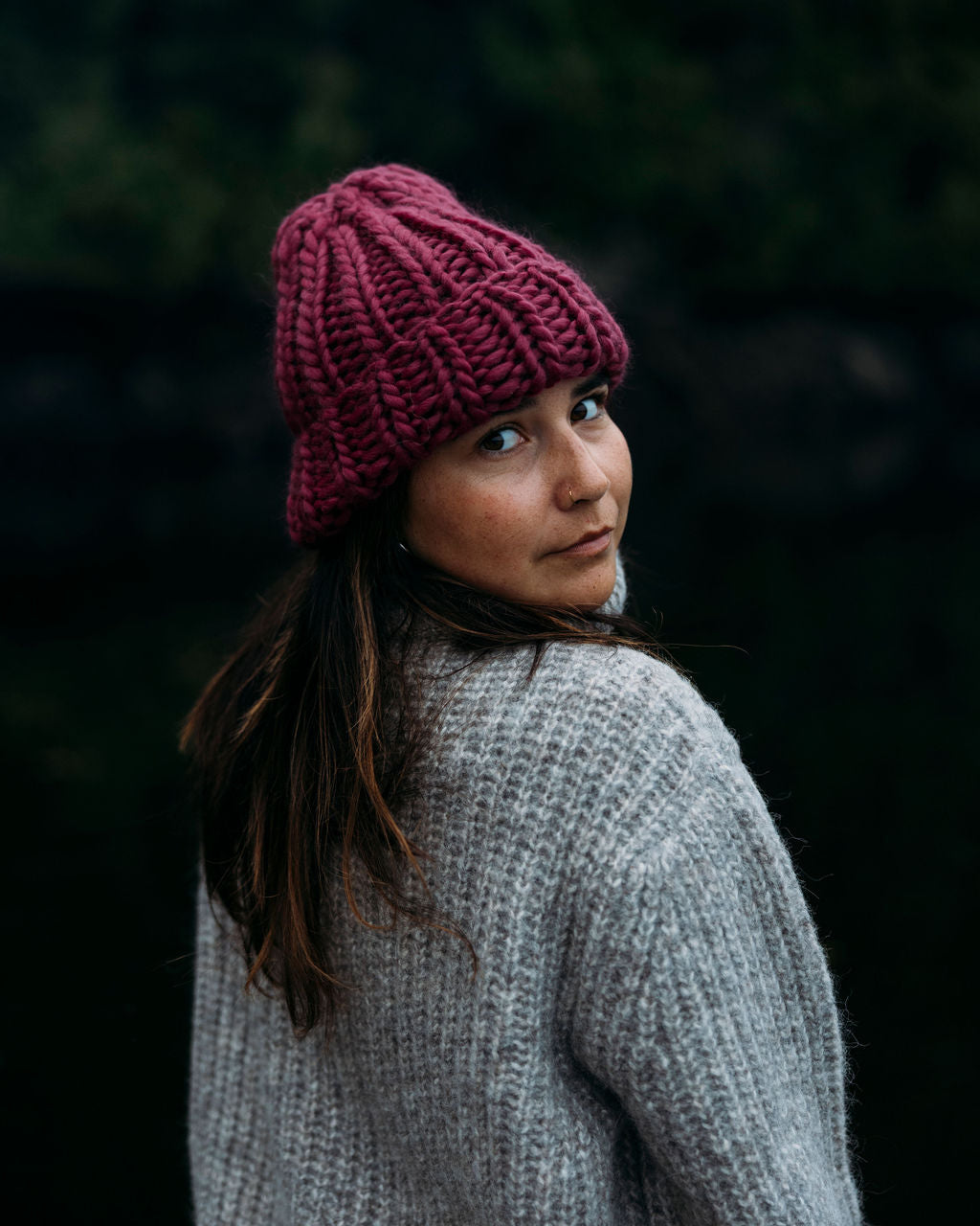 Person wearing a Magenta Diane Rudge knit toque and grey Bare Knitwear sweater standing against a forest background