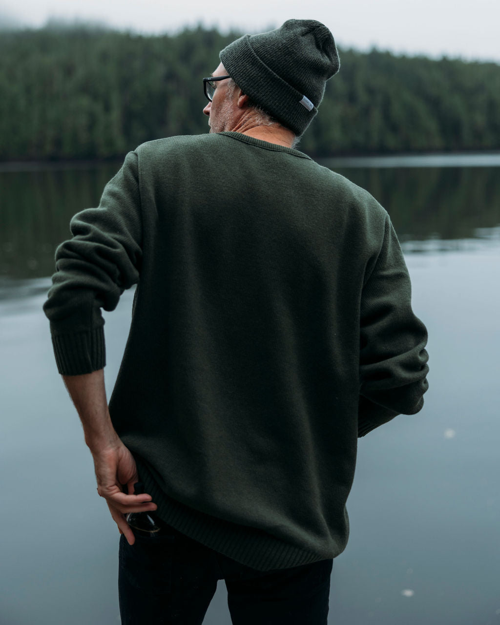 Man wearing a green anian fisherman's sweater and beanie standing by a body of water with trees in the background