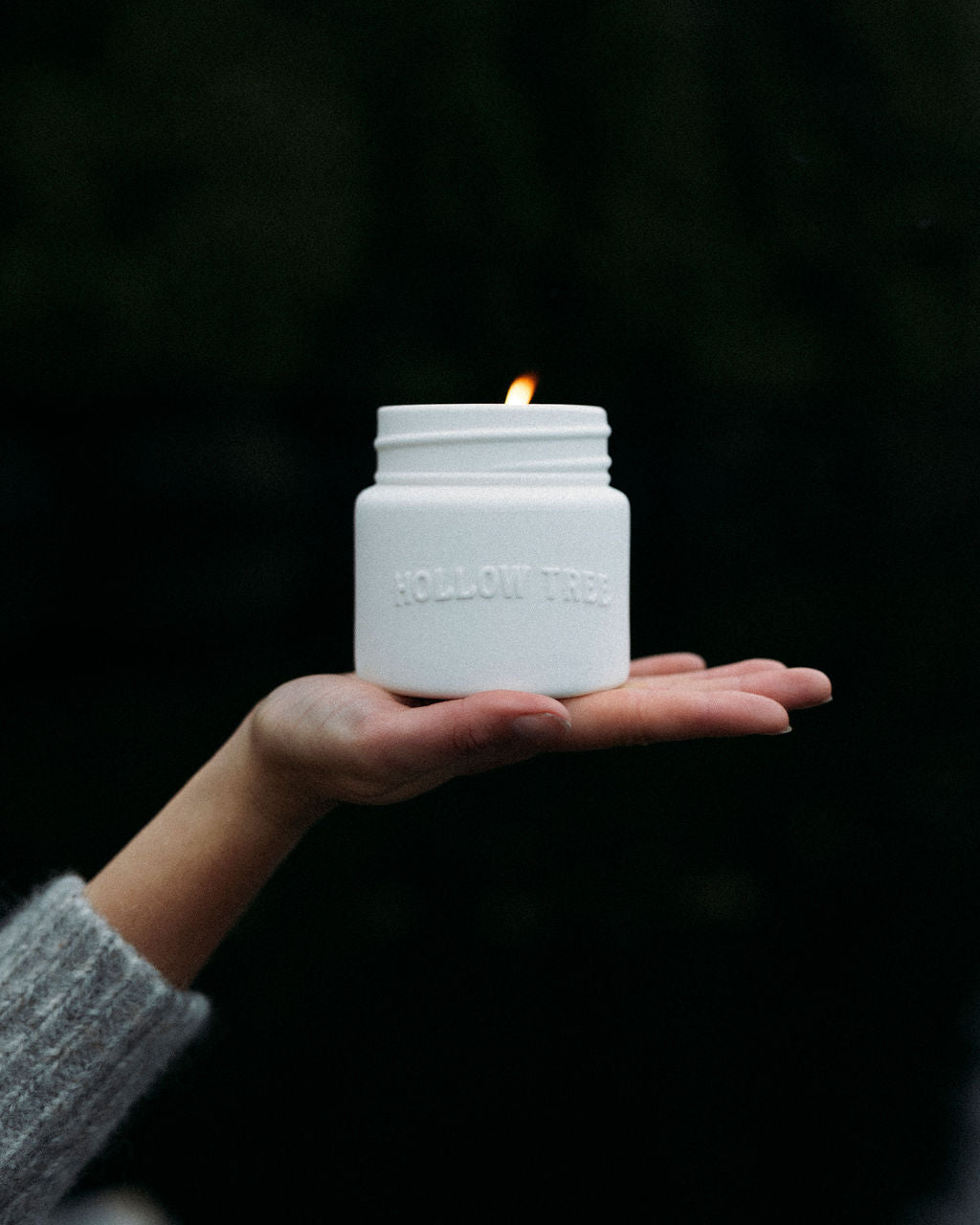 Hand holding a white Hollow Tree Candles jar with a flame against a dark background