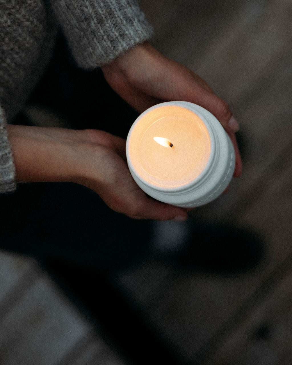 Person holding a small lit Hollow tTee Candle in a white container on a wooden floor.