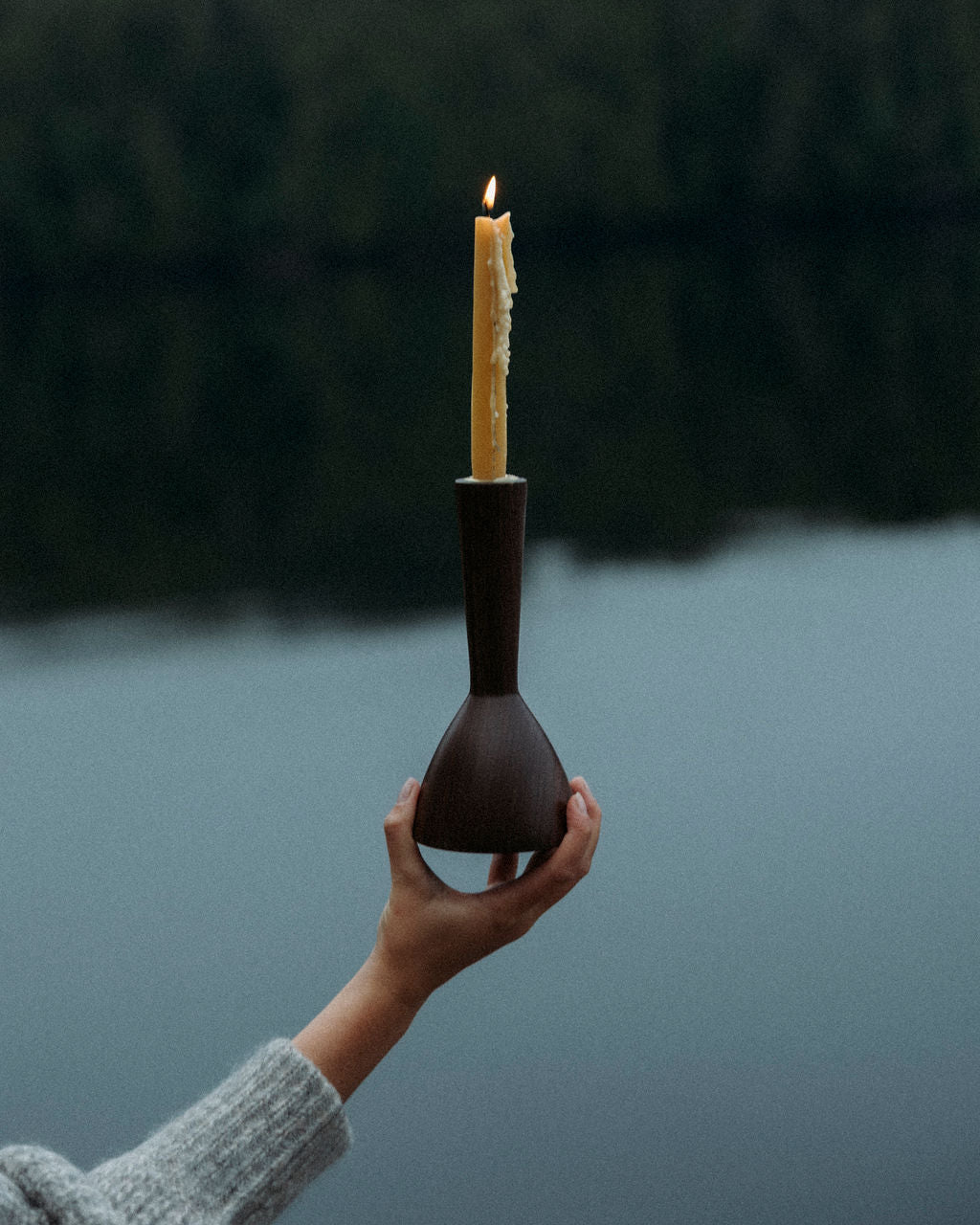 Hand holding a walnut wooden Miller + Co. Wood candle holder with a lit Crowfoot collective beeswax candle against a blurred natural background