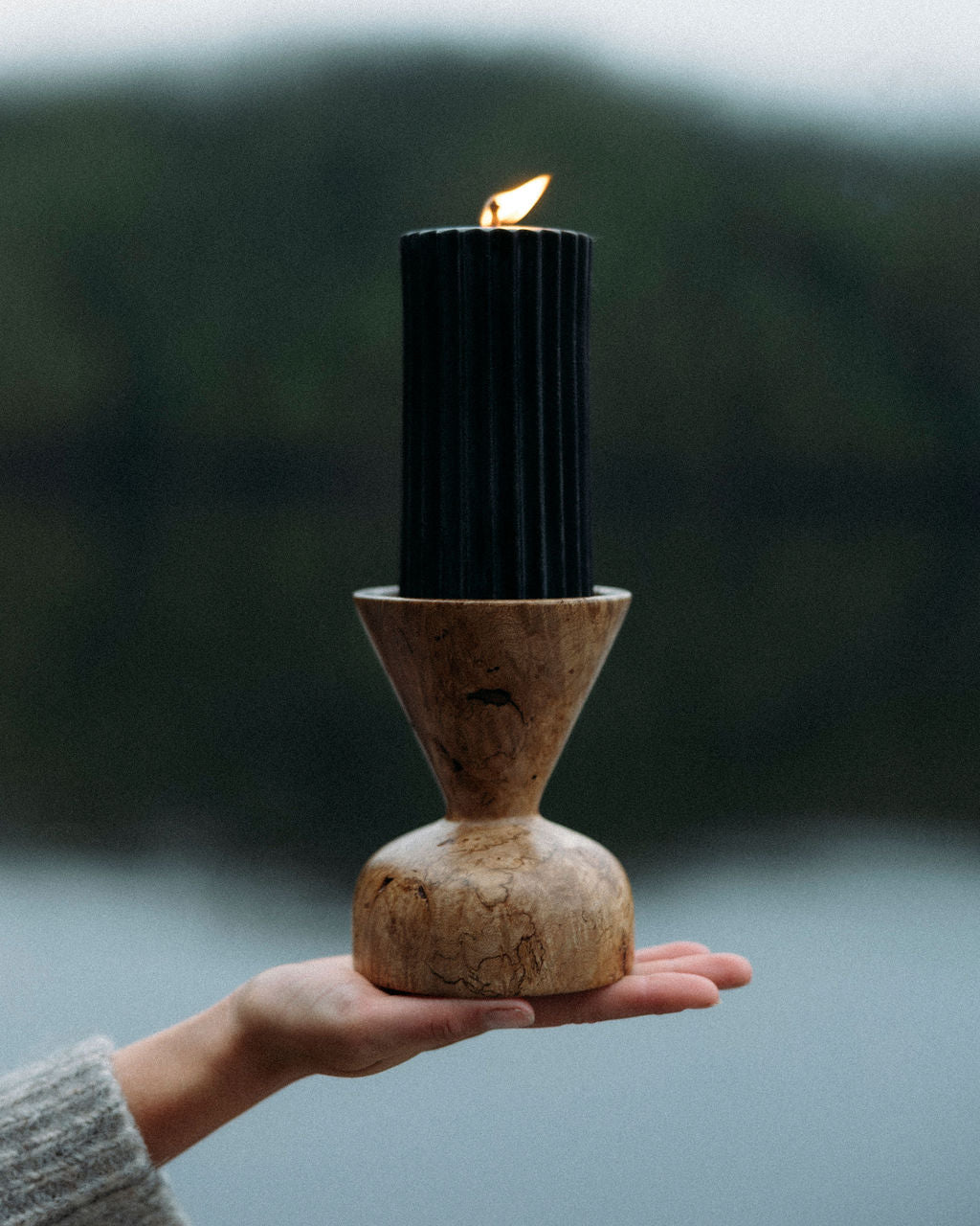 Hand holding a rustic candle holder with a lit candle against a blurred natural background