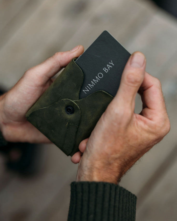 Hand holding a green leather case with 'Nimmo Bay' branding against a blurred background