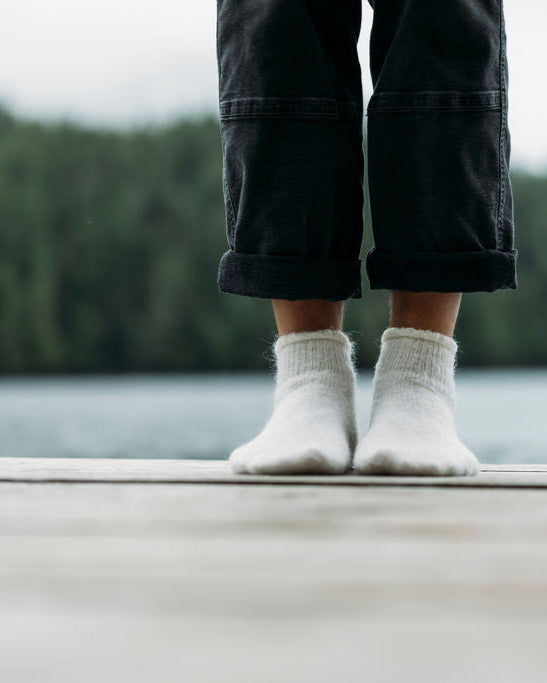 Person wearing white thermohair ankle socks socks and dark pants standing on a dock with a forest in the background