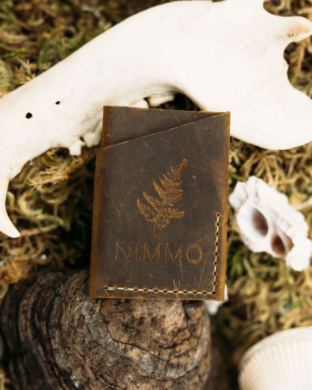 Brown leather wallet with 'NIMMO' branding on a natural background with antlers and moss.