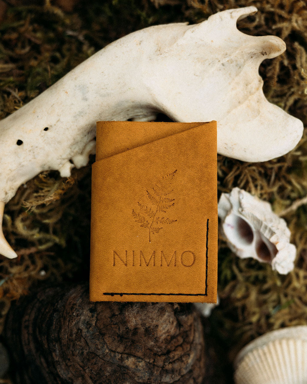 Brown leather notebook with 'NIMMO' branding on a natural background with shells and a bone.