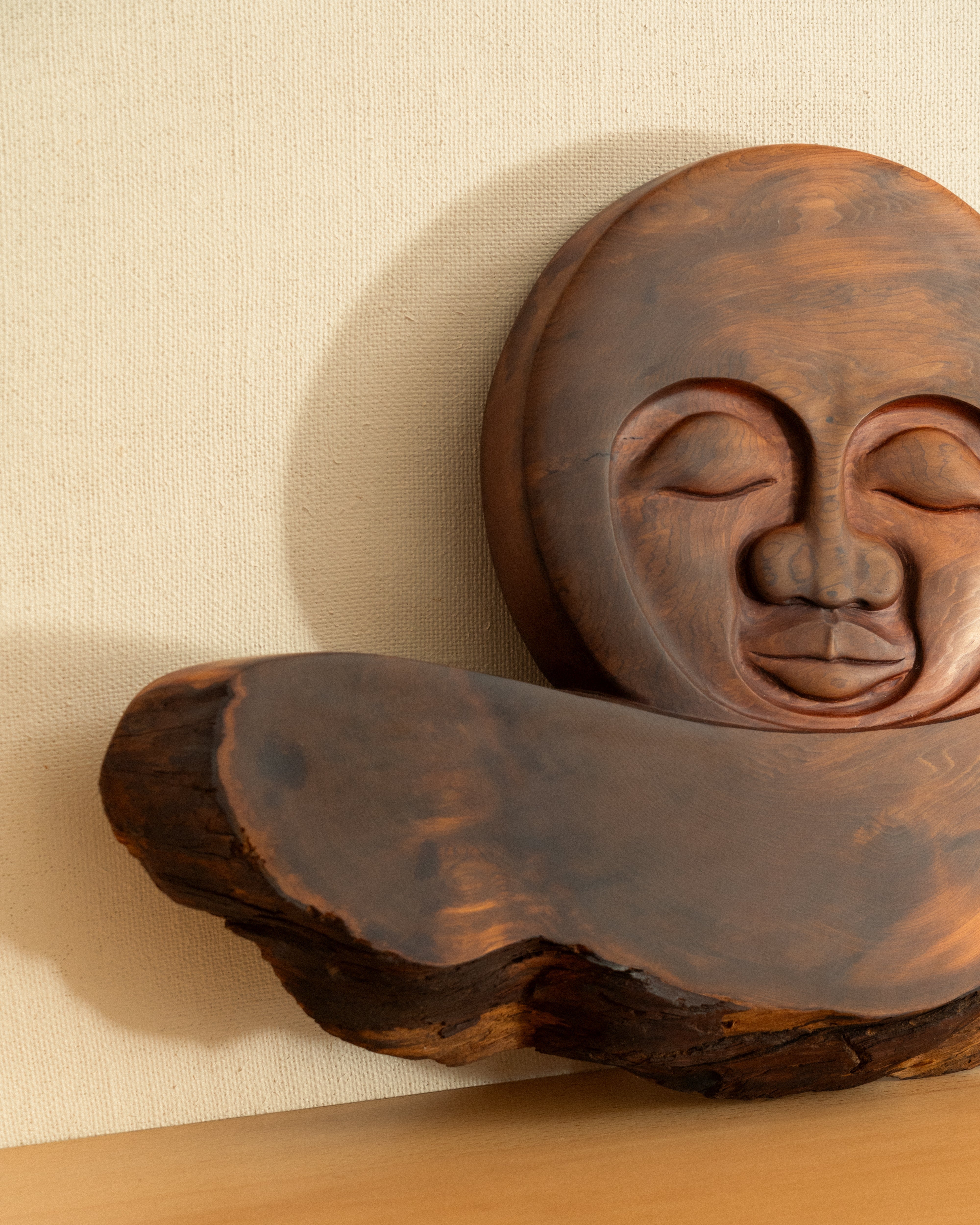 Wooden carving of a face with closed eyes on a wooden shelf against a beige wall.