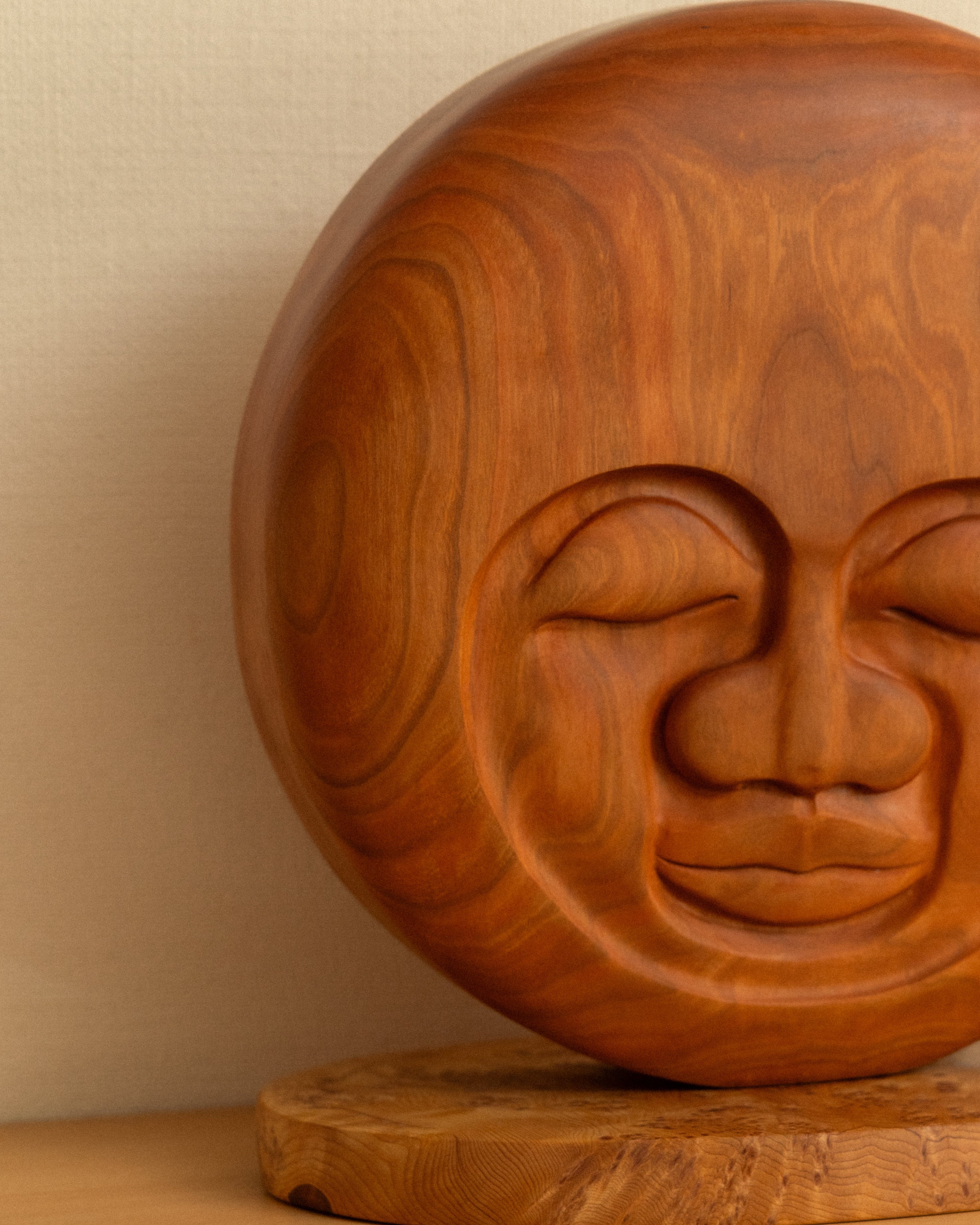 Wooden sculpture of a moon with a serene expression on a wooden base against a beige wall.