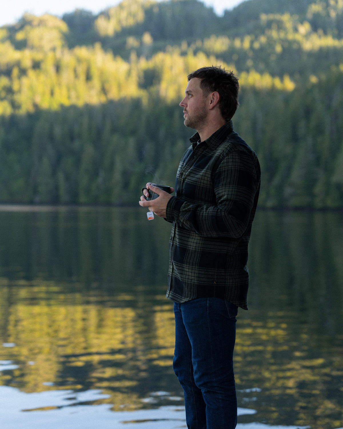 A man wearing a dark green and black plaid flannel shirt standing outdoors on a wooden dock with a blurred forest background and ocean background
