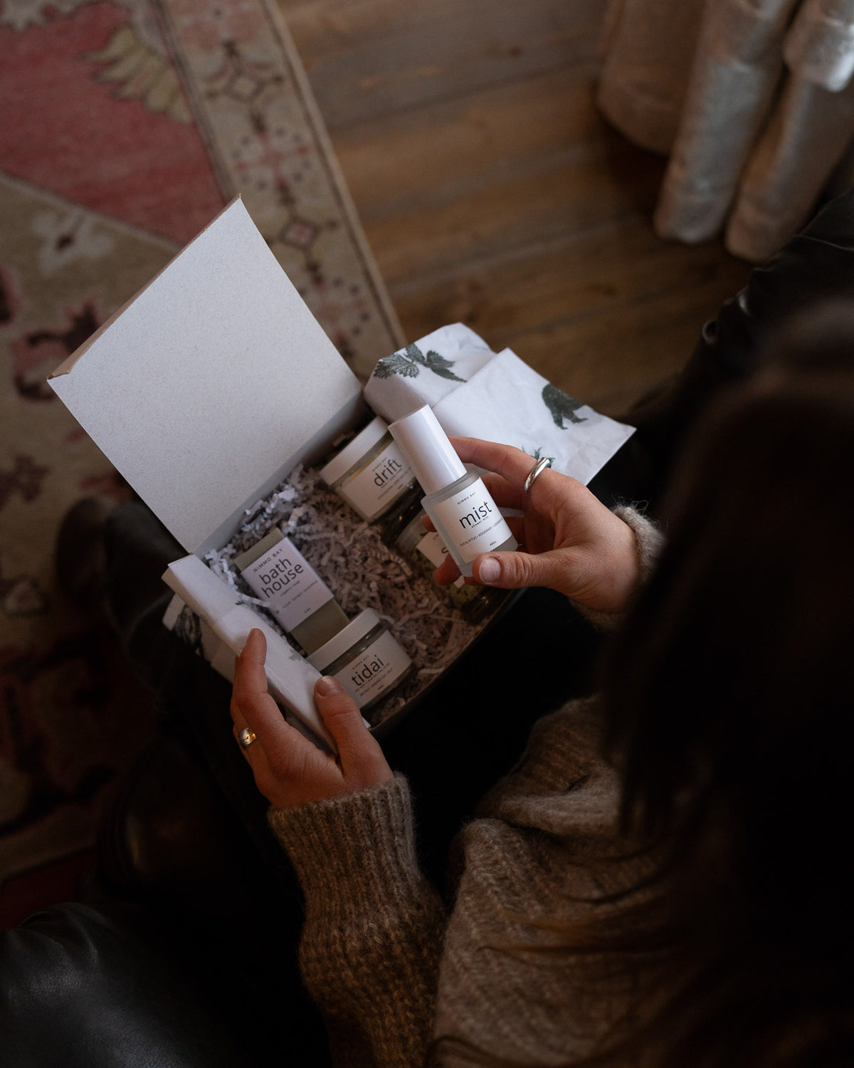 A person holding an open box of skincare products, including a white bottle labeled 'mist' and several cream jars, in a cozy indoor setting with a patterned rug and wooden floor in the background.