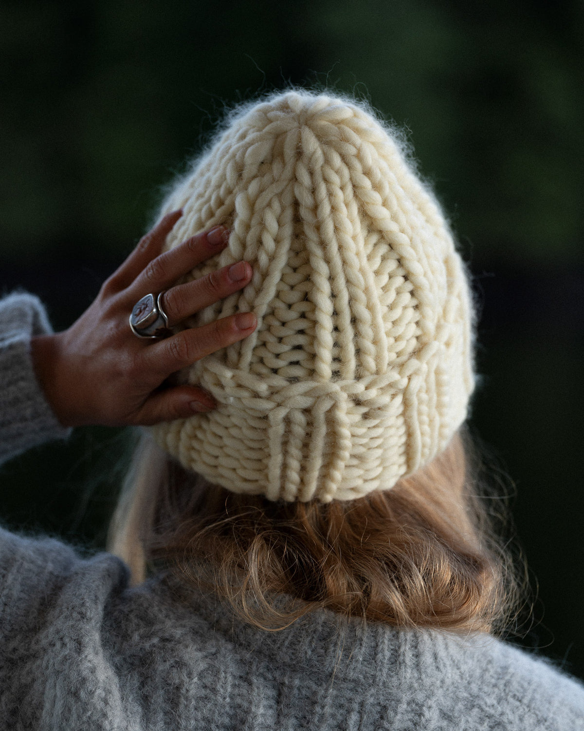 a woman from behind wearing a toque and knit sweater against a background of trees