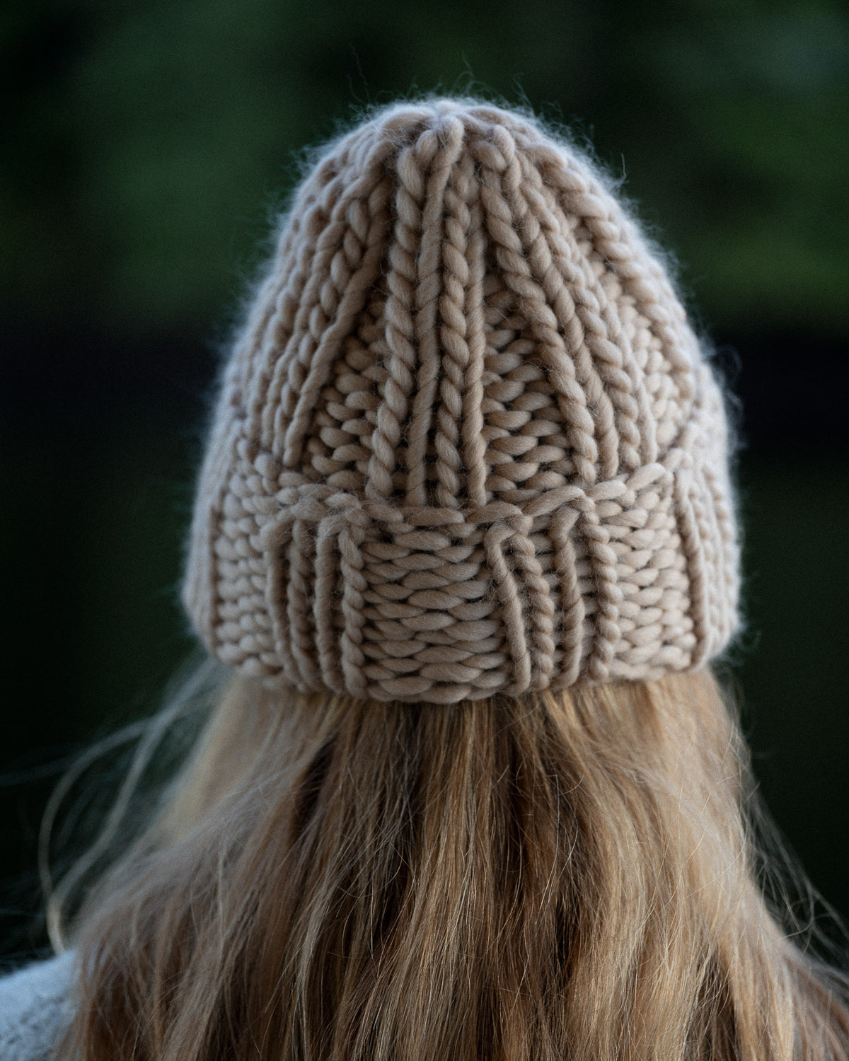 a woman from behind wearing a toque and knit sweater against a background of trees