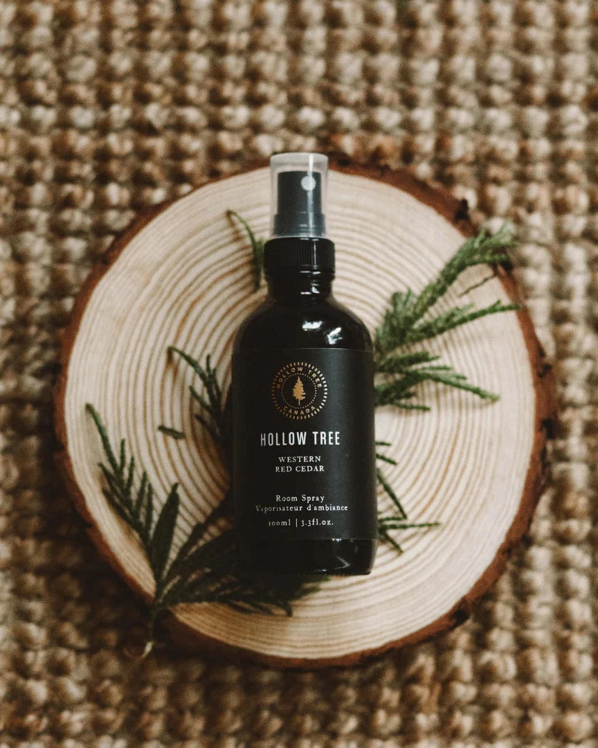 A bottle of Hollow Tree Natural Red Cedar room spray sits on a round wood slice, styled with green sprigs on a textured woven jute background.