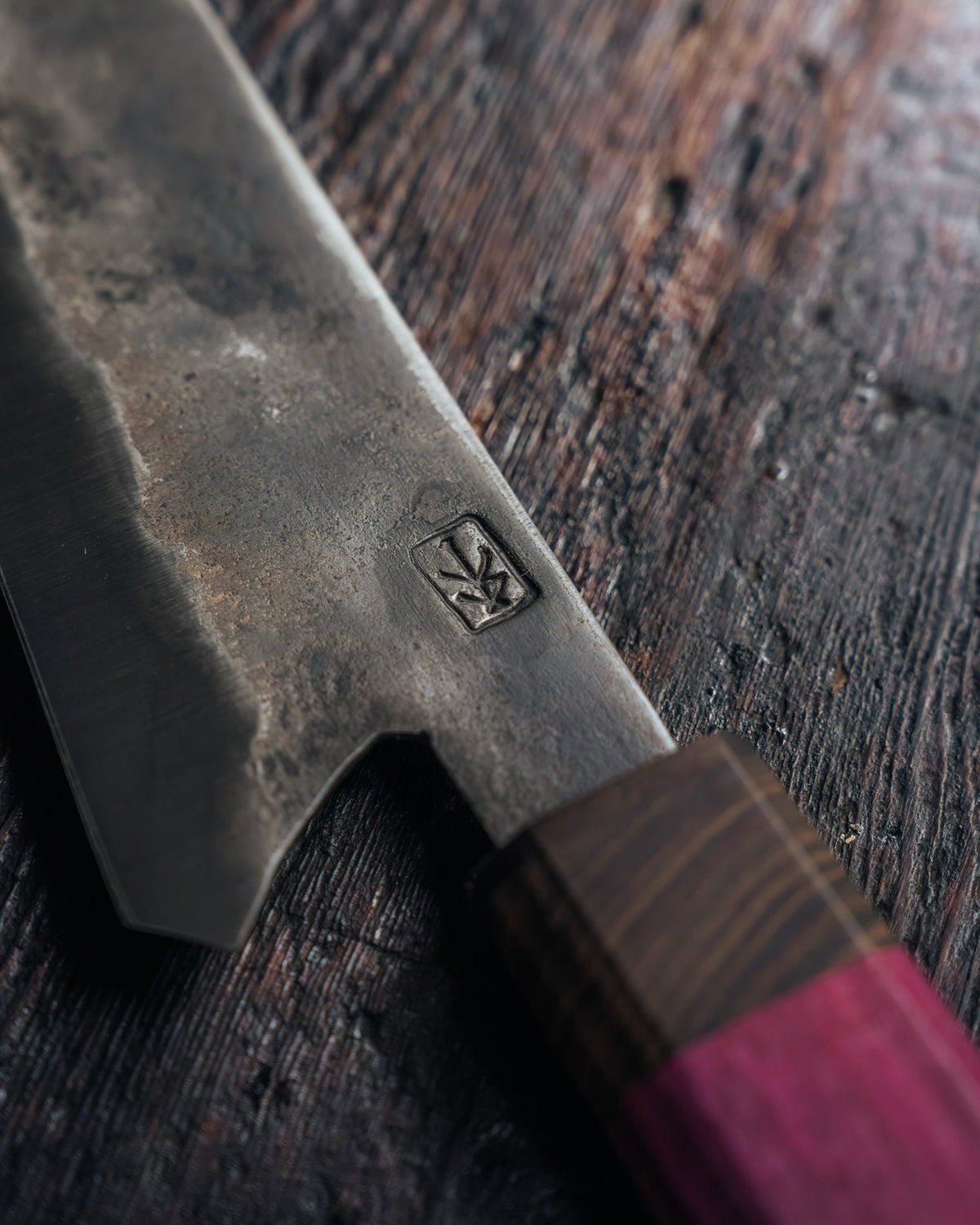 A close-up of a Japanese-style kitchen knife with a dark, textured steel blade featuring a stamped logo, resting on a rustic dark wood surface. The handle is made of dark wood with a vibrant magenta-pink wrapped section.