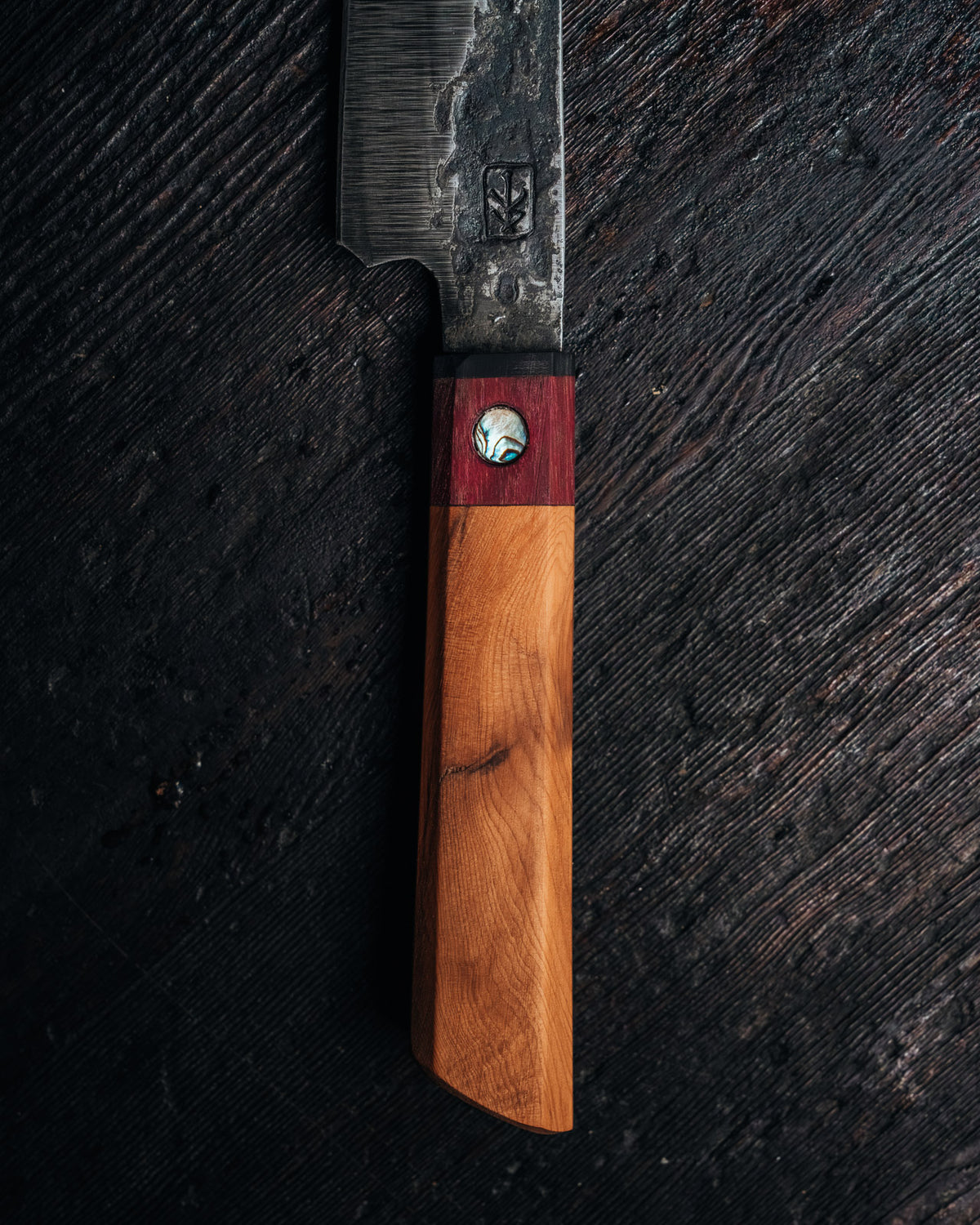 A close-up of a handcrafted knife with a dark textured steel blade and a dual-tone wooden handle, placed diagonally on a dark wood background.