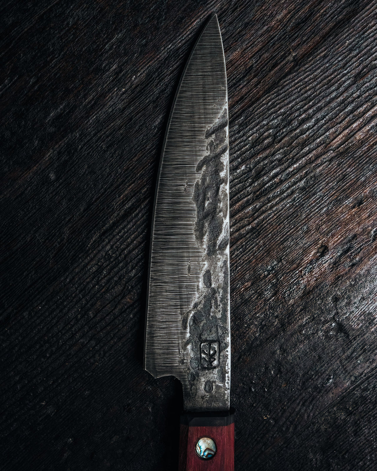 A single chef's knife with a textured, patterned steel blade and a reddish-brown wooden handle, laid vertically on a dark, rustic wood surface.