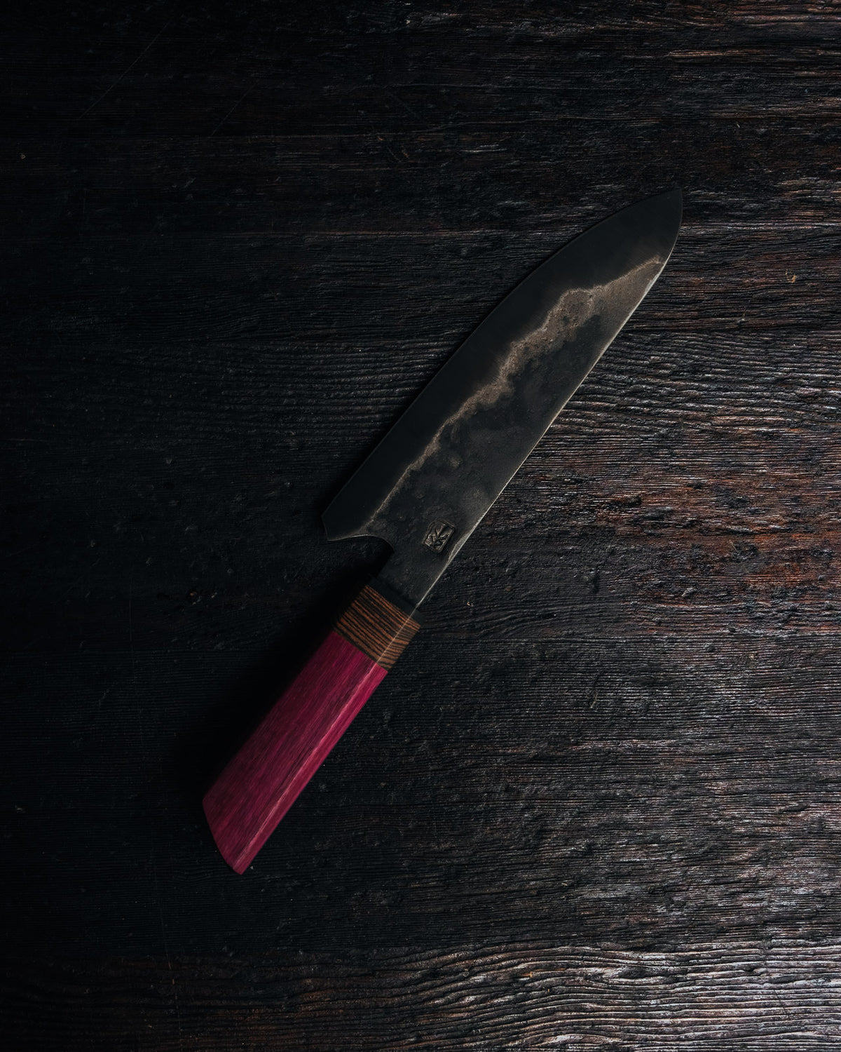 A Japanese-style chef's knife with a dark, textured steel blade and a reddish-pink wooden handle wrapped with a thin brown cord, resting diagonally on a dark, rustic wooden surface.