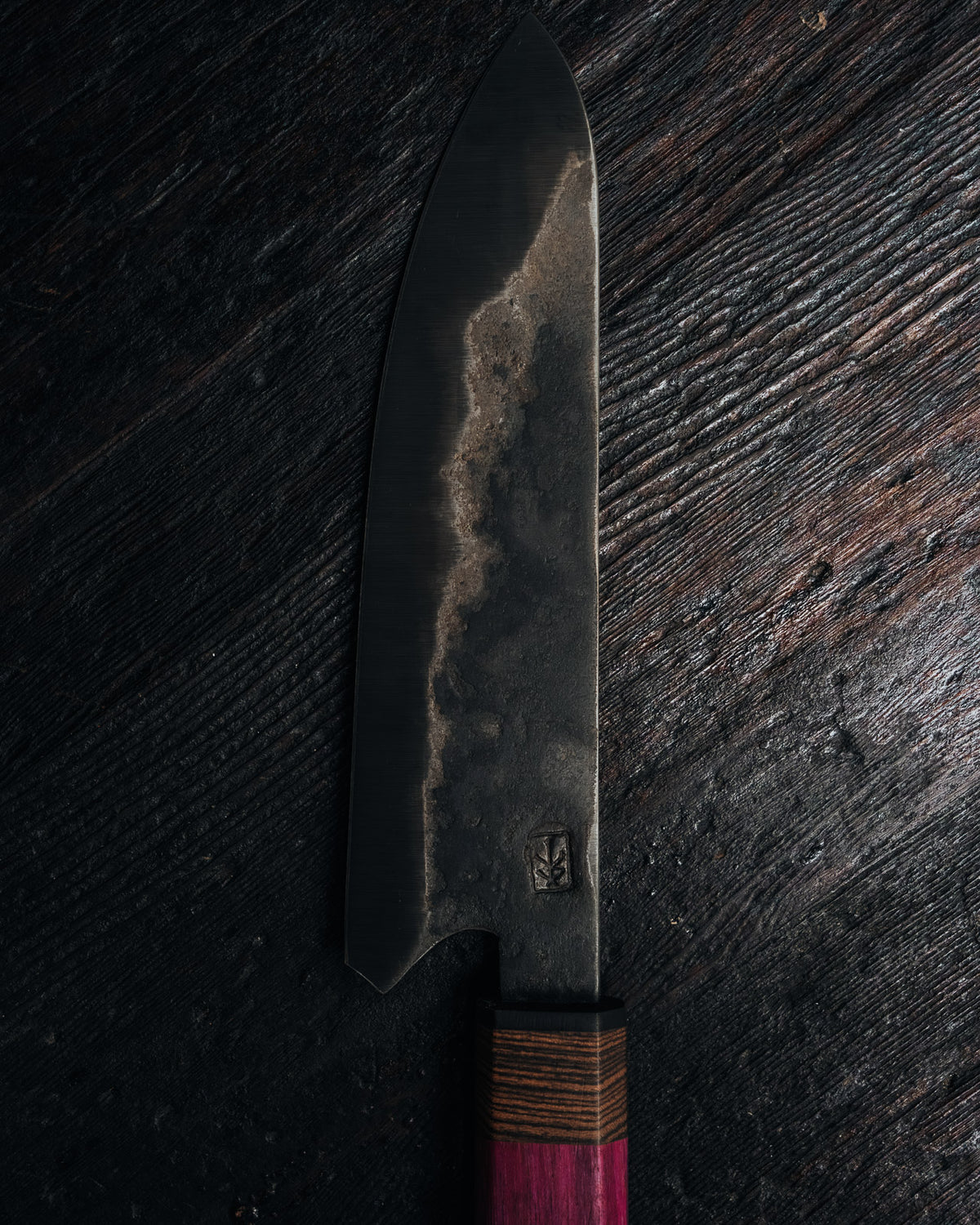A Japanese-style chef's knife with a dark, textured forged steel blade and a reddish-brown wooden handle, displayed vertically on a dark rustic wooden surface.