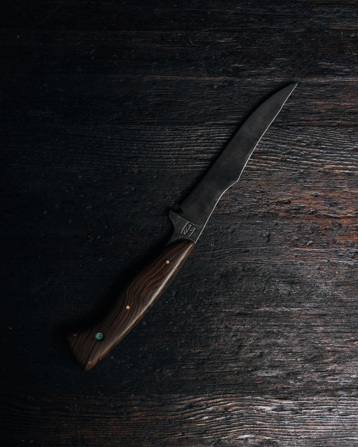 A fixed-blade knife with a dark steel blade and a brown wood-grain handle, placed diagonally on a dark rustic wooden surface.