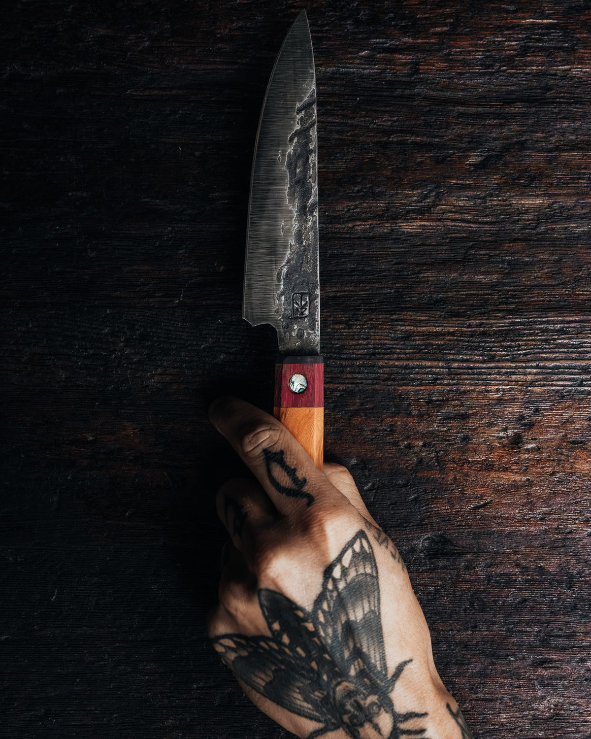 A hand with black ink tattoos holds a fixed-blade knife with a dark textured steel blade, a reddish-brown handle, and a silver bolster against a dark rustic wooden background.