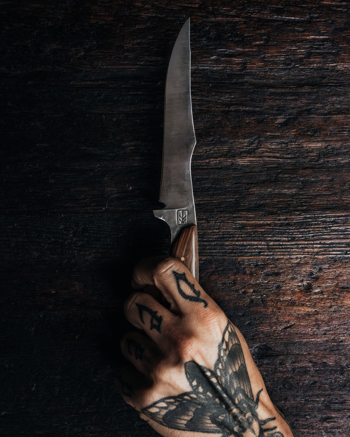 A hand with black tattoos holds a fixed-blade knife with a satin-finished steel blade and a brown wooden handle against a dark, rustic wooden background.
