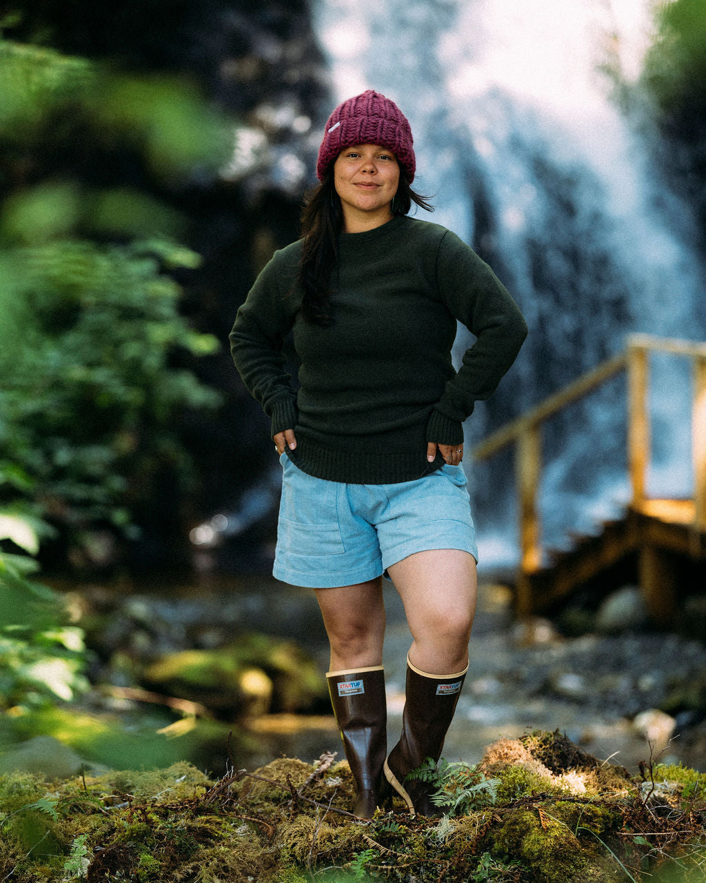 Woman standing in a forest wearing the Anian green frishermans sweater with a waterfall in the background