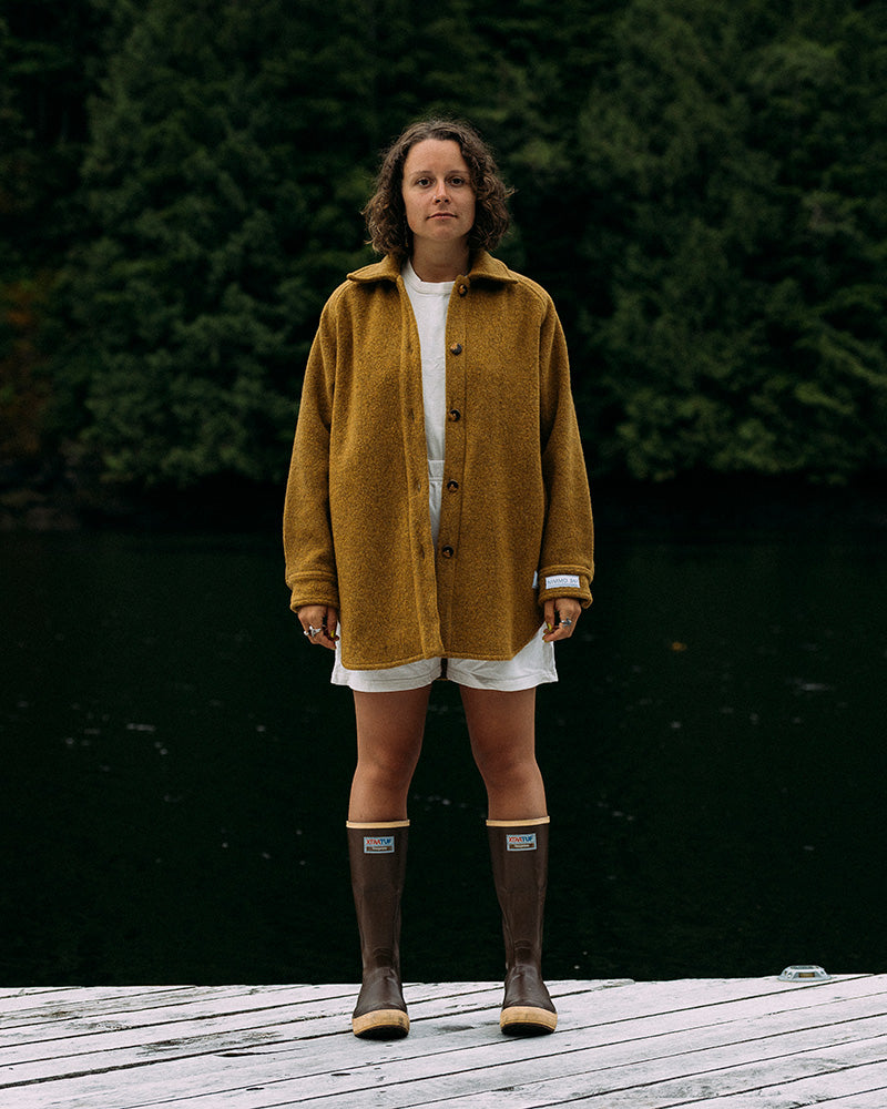 Person wearing a mustard yellow jacket and brown boots standing on a wooden deck with a dark green background.