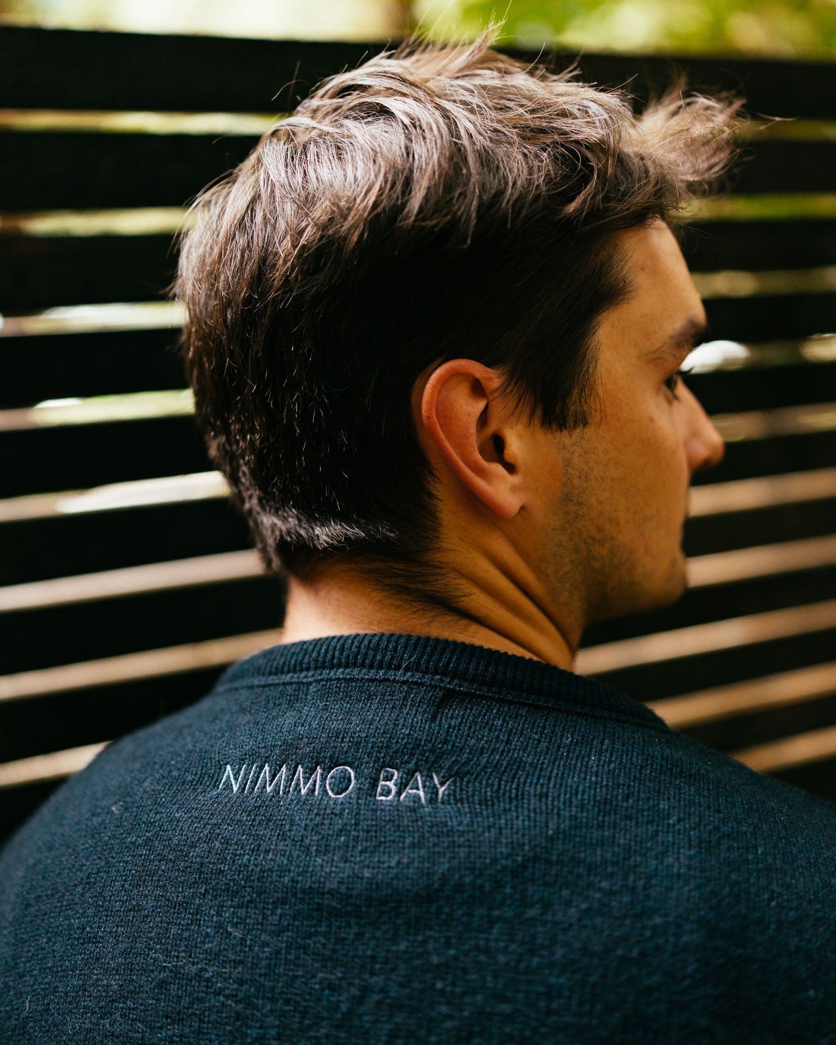 Man wearing a dark blue sweater with 'Nimmo Bay' branding, standing against a blurred natural background.