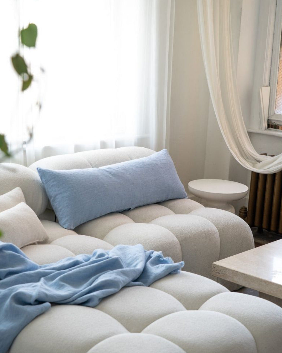 Beige sofa with blue cushions and a blanket in a room with white curtains and a plant.
