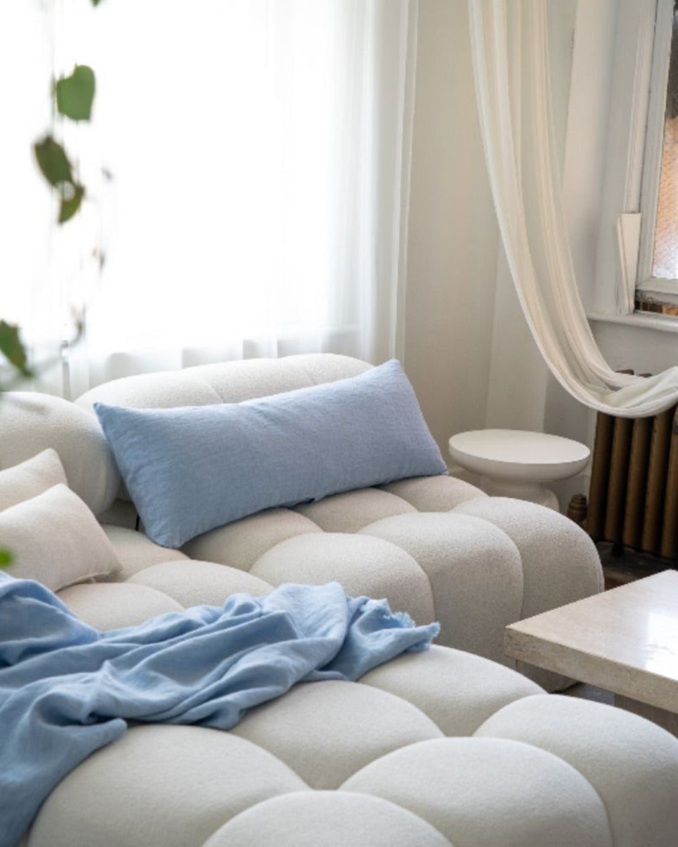 Beige sofa with blue cushions and a blanket in a room with white curtains and a plant.