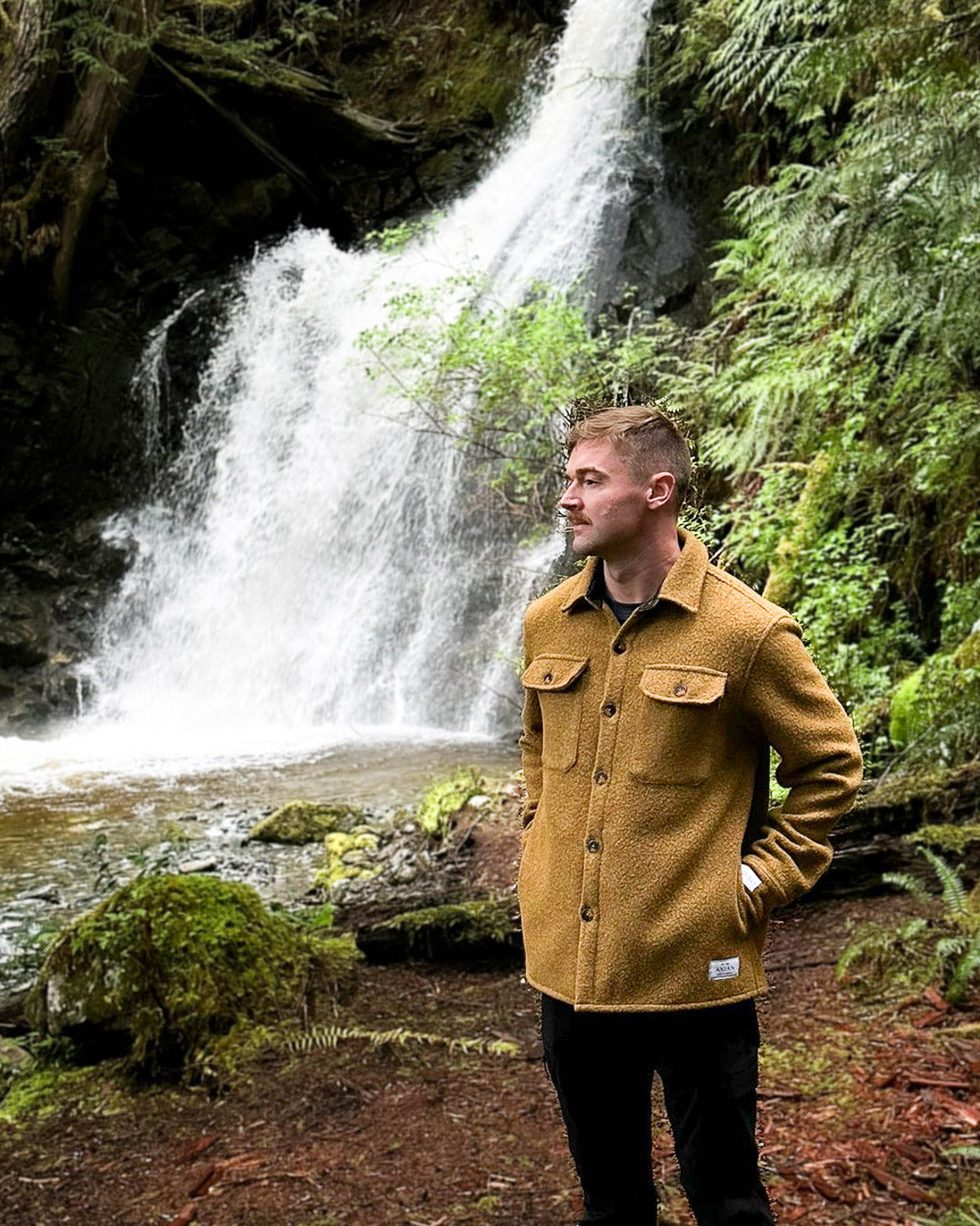 man standing in front of waterfall with gold anian field coat at Nimmo Bay
