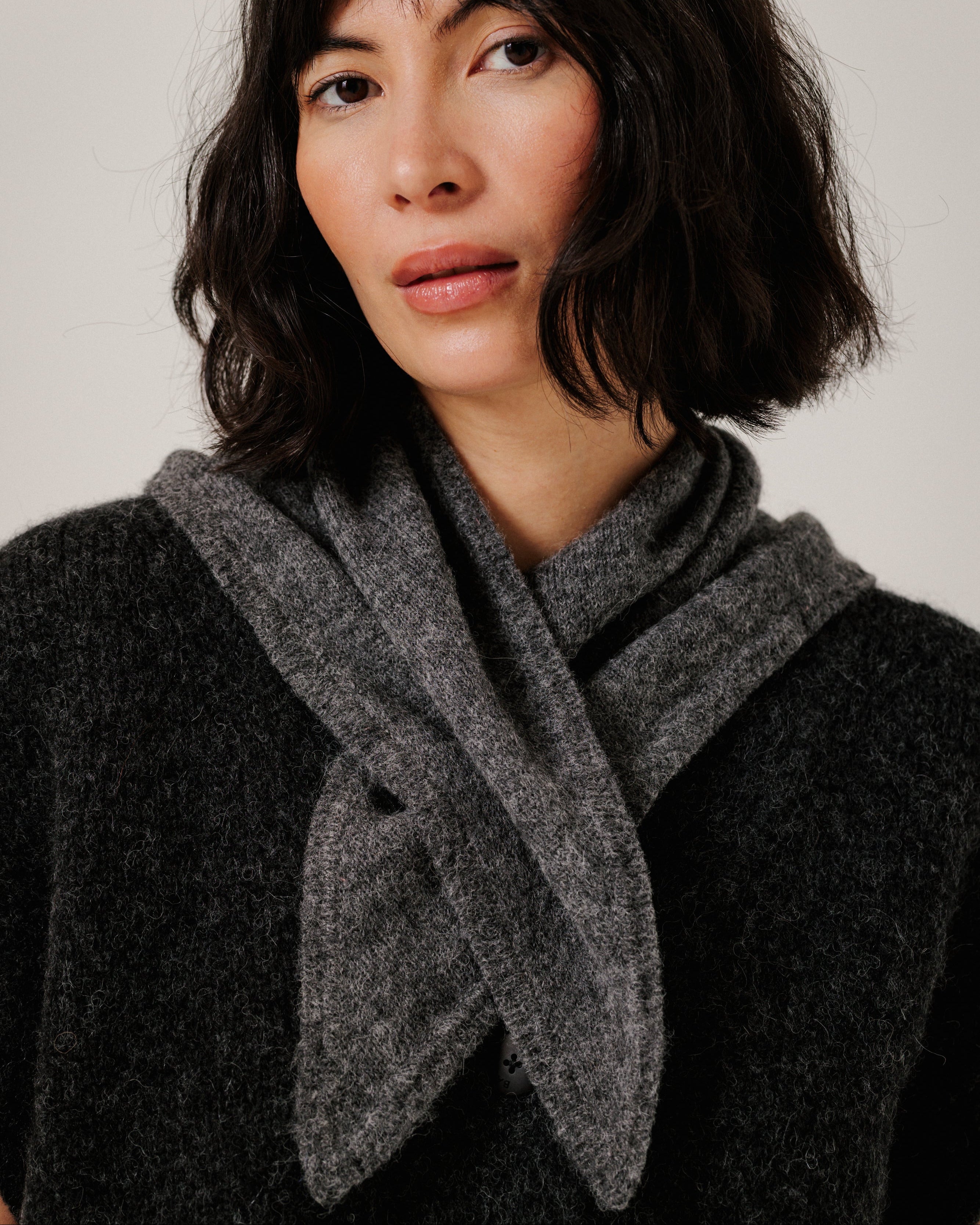 A woman with dark shoulder-length hair wearing a dark charcoal knit sweater and a thick, textured gray knit scarf wrapped around her neck, photographed against a plain light gray background.
