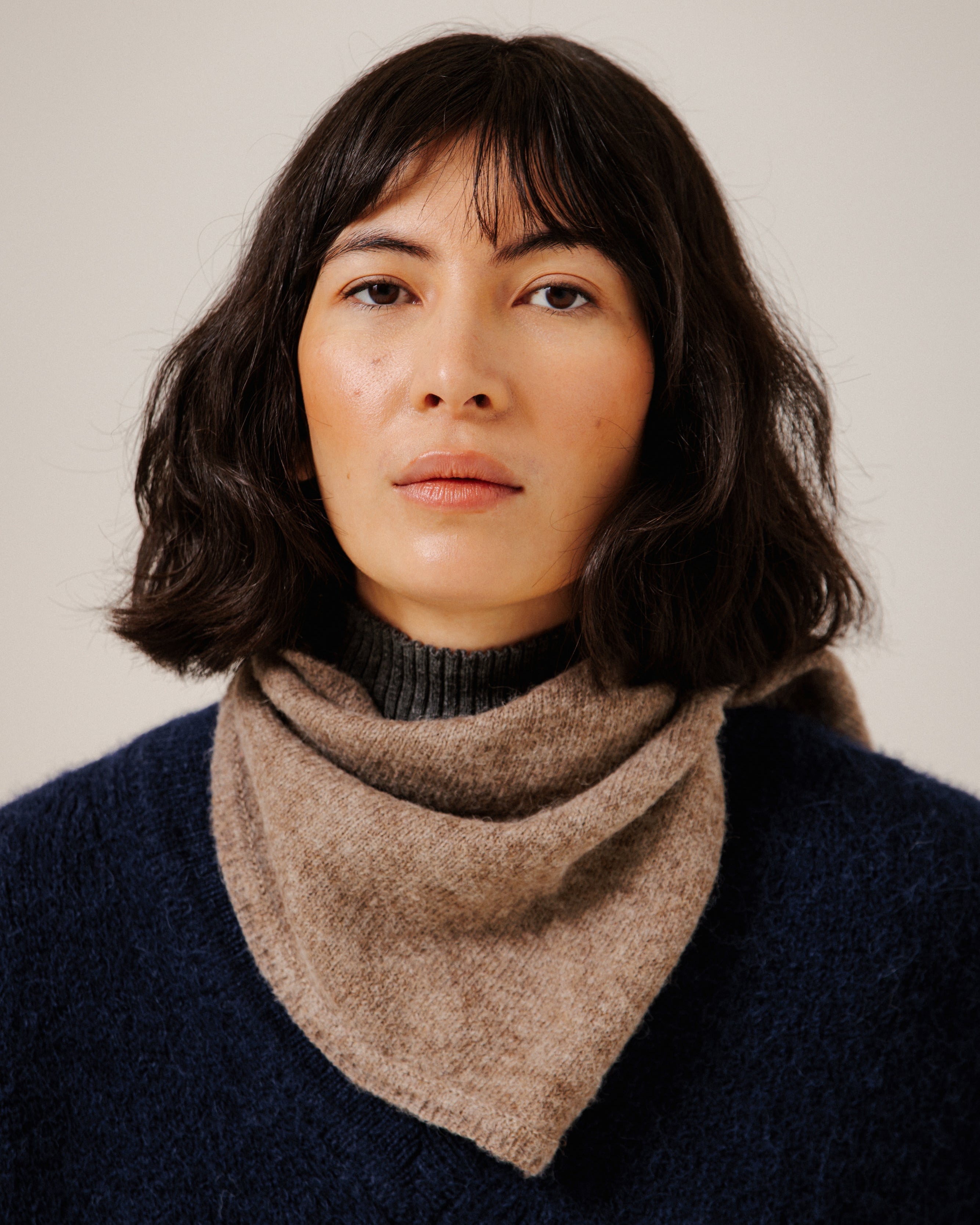A woman with dark shoulder-length hair wearing a soft taupe-brown fuzzy cowl neck warmer over a dark ribbed turtleneck sweater, set against a plain light beige background.