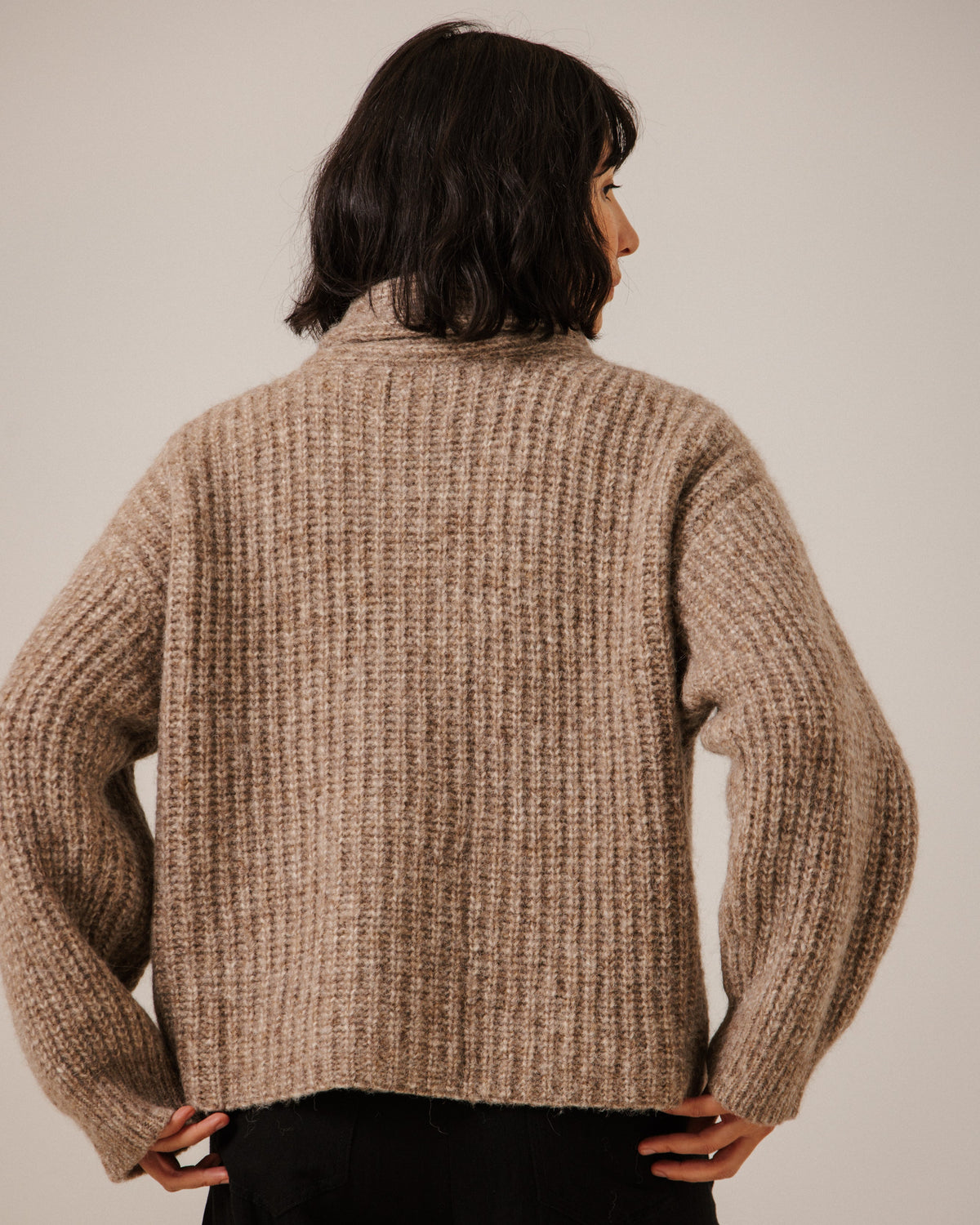 A person shown from the back wearing a chunky ribbed knit sweater in a warm taupe-beige color, paired with black pants, against a plain light beige studio background.
